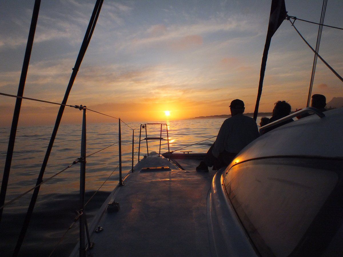 Silhouetted individuals are seated on a sailboat's deck, watching a vibrant sunset over calm waters. The sky is painted with warm hues, and the sun is just above the horizon. The boat's rigging and structure frame the peaceful ocean scene, creating a serene atmosphere.