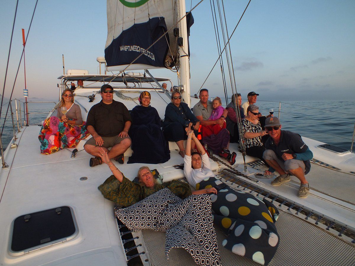 A group of people sitting and lying on the deck of a yacht, covered with colorful blankets. The sail is visible above, and the ocean and sky provide a serene backdrop. The atmosphere is relaxed, suggesting a leisurely outing at sea.