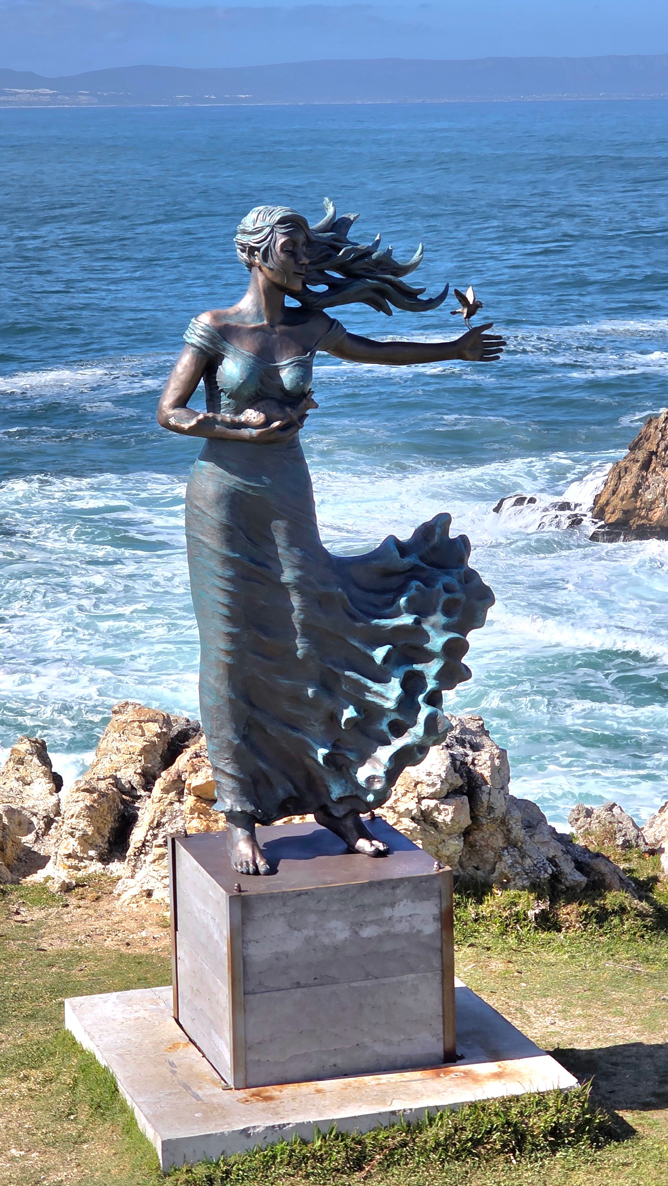 A dynamic bronze sculpture of a woman with flowing hair and dress, positioned on a rocky coastline. The figure appears to embrace the ocean breeze, with waves crashing in the background, symbolizing freedom and connection to nature in the Hermanus art scene.
