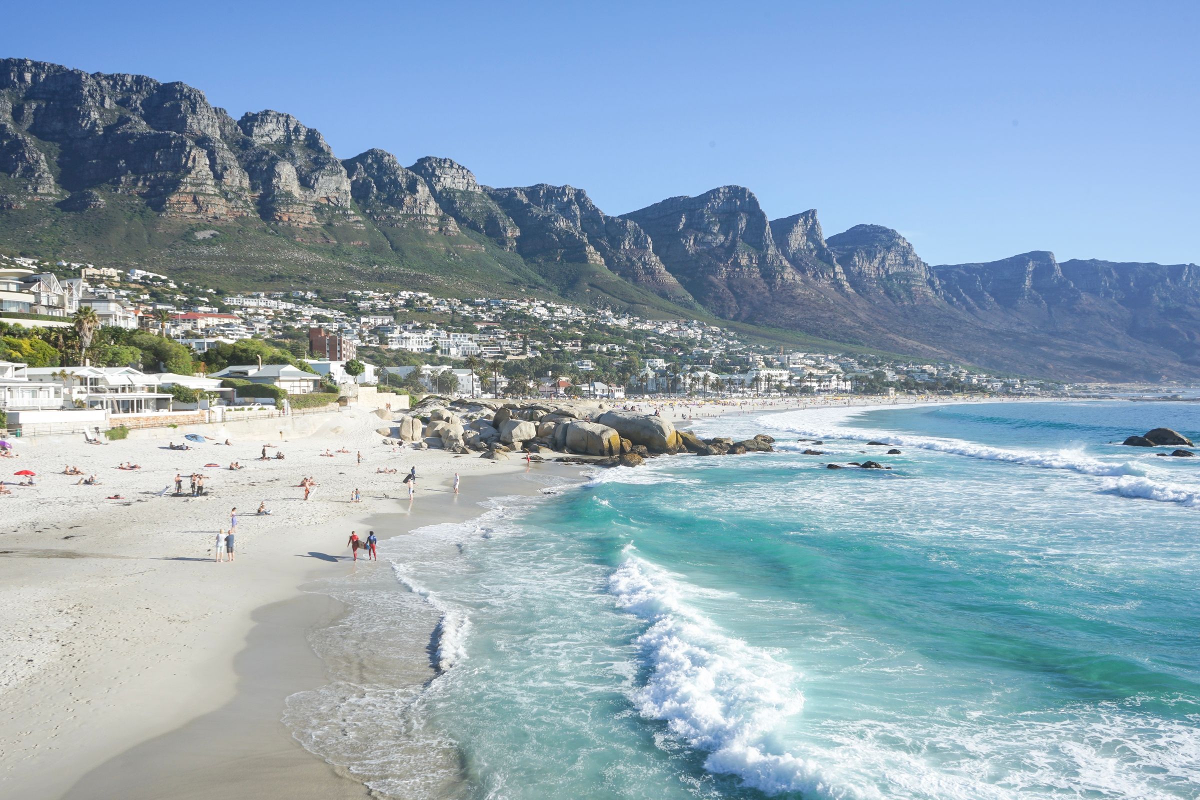 A stunning view of a Cape Town beach featuring turquoise Atlantic waves crashing onto white sand. The scenic backdrop includes the majestic Twelve Apostles mountain range and a vibrant beachside community under a clear blue sky, showcasing the city's natural beauty and lively atmosphere.