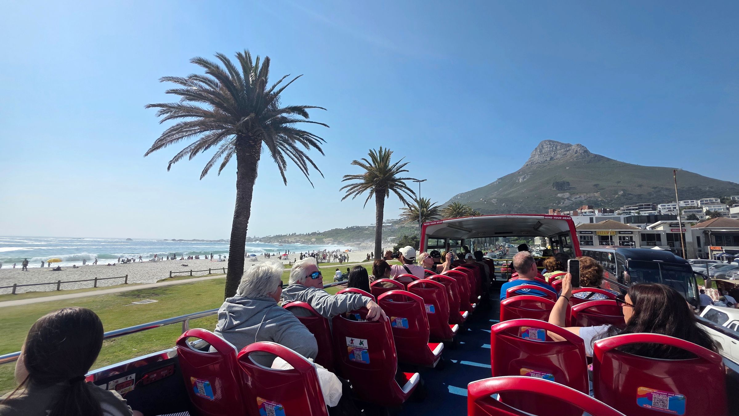 Passengers enjoy views from a red open-top bus in Cape Town. The bus travels along a coastal route with palm trees and a mountain, offering a scenic exploration of landmarks and beaches.