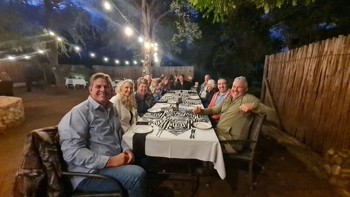 A group of people seated around a long table outdoors, enjoying a dinner under string lights. The table is set with patterned tablecloths and dinnerware. Trees and a wooden fence surround the area, creating a cozy evening atmosphere.