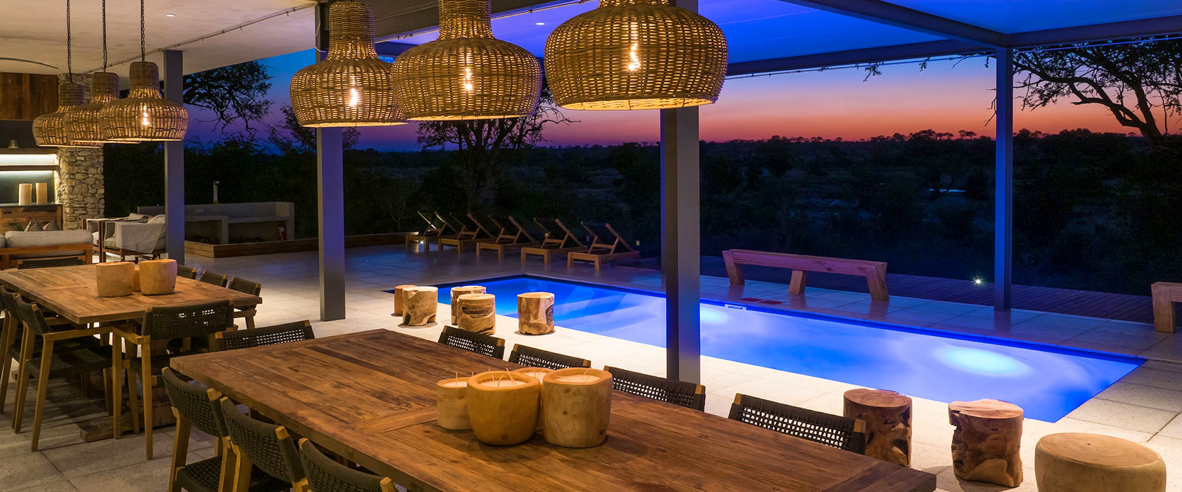 Outdoor dining area at Kruger Safari Lodge featuring wooden tables and chairs, wicker pendant lights, and a glowing pool. The setting overlooks a scenic twilight view, blending luxury and nature in the African wilderness.