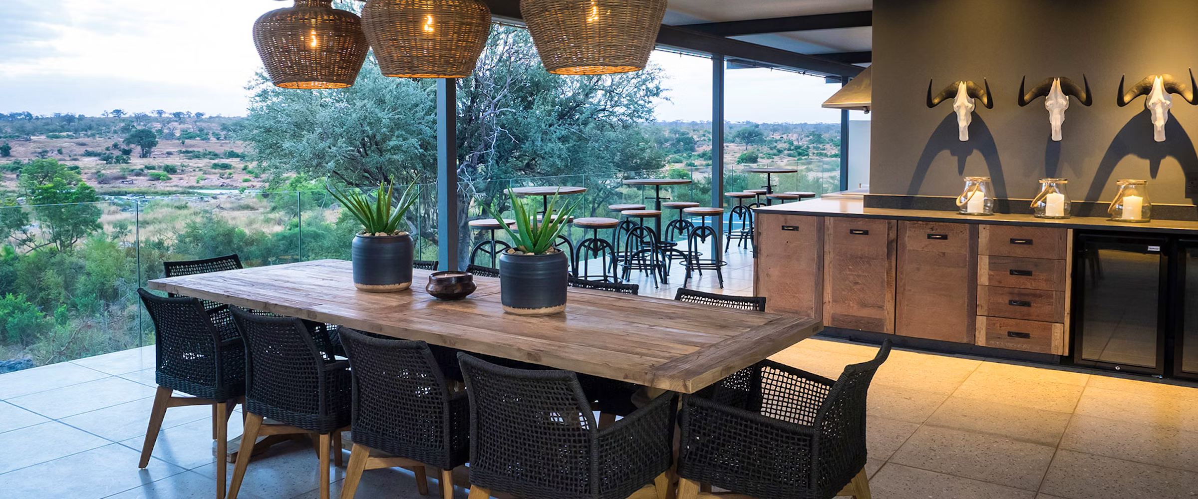 An elegant outdoor dining space with a large wooden table and woven chairs. Overlooking a natural landscape, the area features hanging wicker lights, decorative plants, and a rustic bar with animal skull decor, blending luxury with nature.