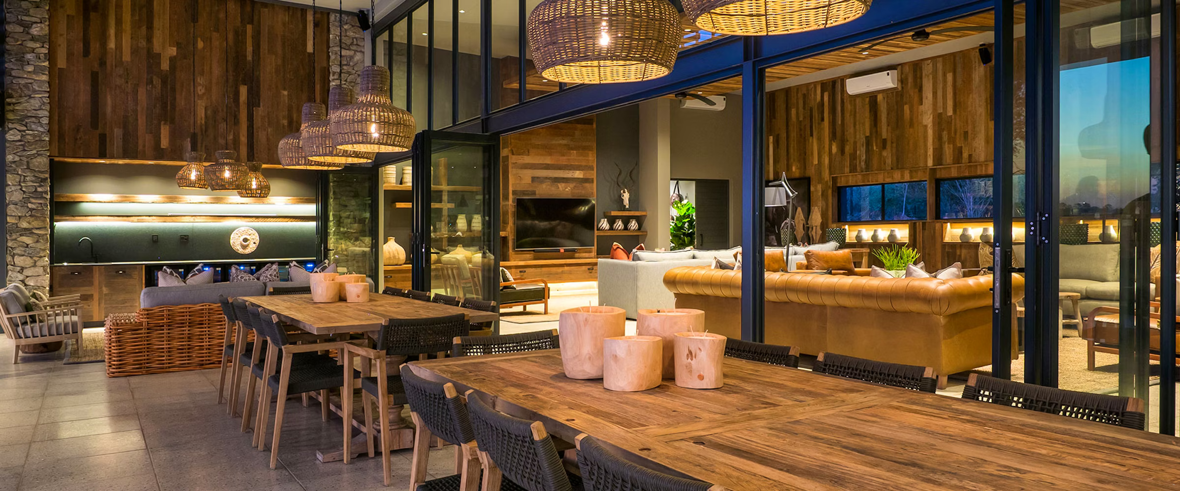 A luxurious space at Kruger Safari Lodge blending indoor and outdoor living. Features rustic wooden tables, wicker chairs, ambient lighting, and plush sofas against stone and wood-paneled walls, creating an inviting atmosphere for dining and relaxation.