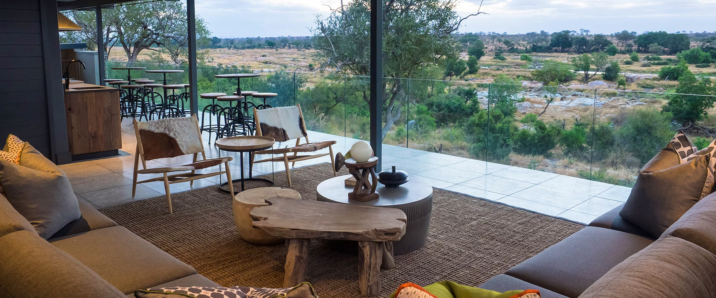 A luxury lounge at Kruger Safari Lodge with modern seating, natural wood tables, and floor-to-ceiling windows offering panoramic views of the African wilderness. The space combines comfort with nature, featuring cowhide chairs, plush cushions, and an inviting atmosphere.