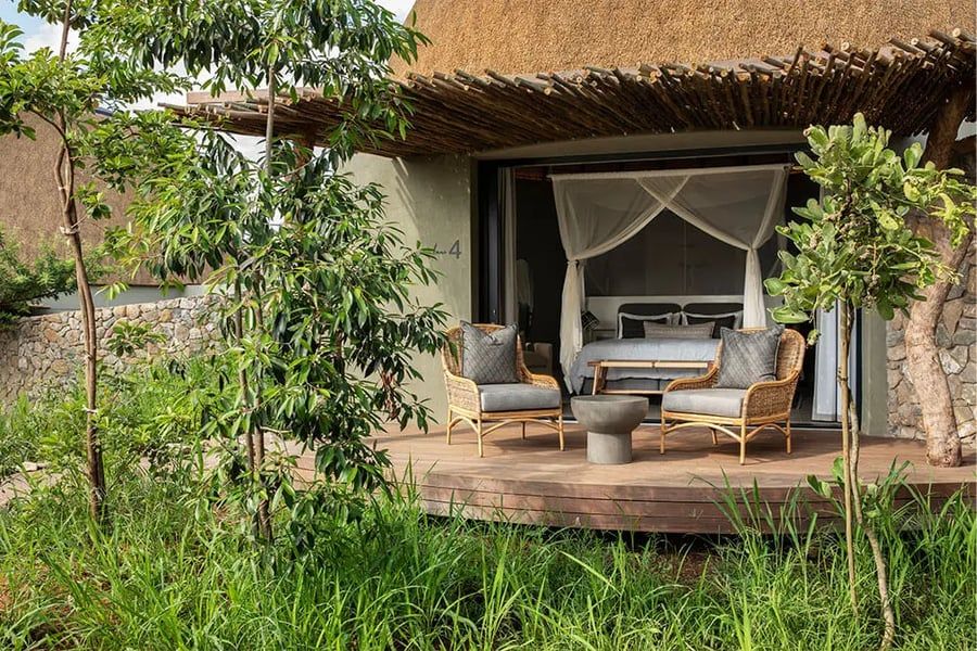 A serene outdoor patio with two wicker chairs and a small table set on a wooden deck. The area is surrounded by lush green plants and trees. A cozy bedroom with a canopy bed is visible through sliding glass doors, creating a tranquil and inviting atmosphere.