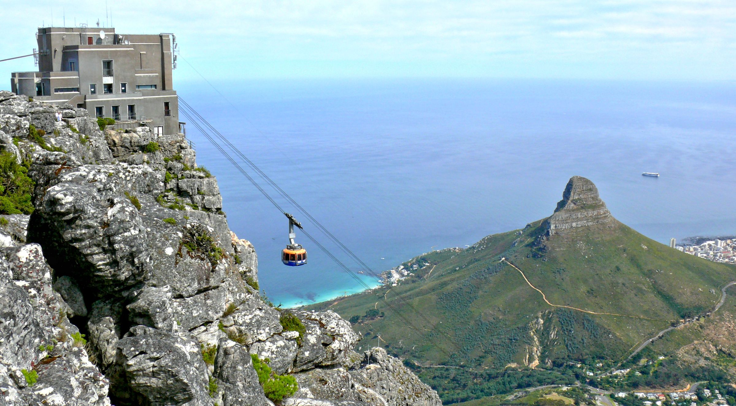 Table Mountain's cable car glides towards the summit, offering sweeping views of the Atlantic Ocean and surrounding landscape. The rugged mountain terrain is prominent, with Lion's Head visible in the background. The scene captures the essence of Cape Town's natural beauty and UNESCO status.