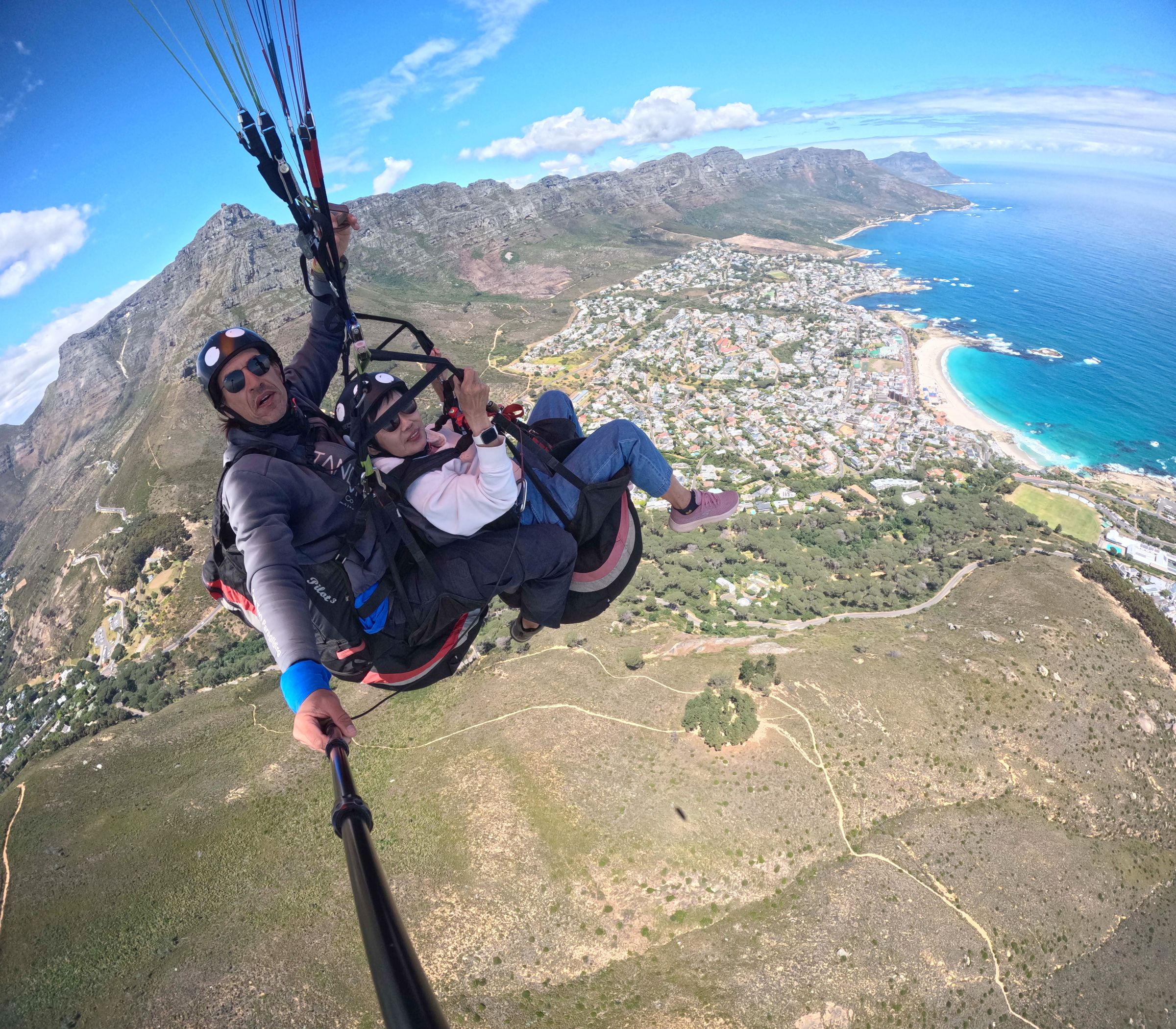 Two people paragliding over a scenic landscape with mountains and coastline. The view includes a sprawling city, flat-topped mountain, and clear blue ocean, highlighting the natural beauty and offering an exhilarating aerial perspective.