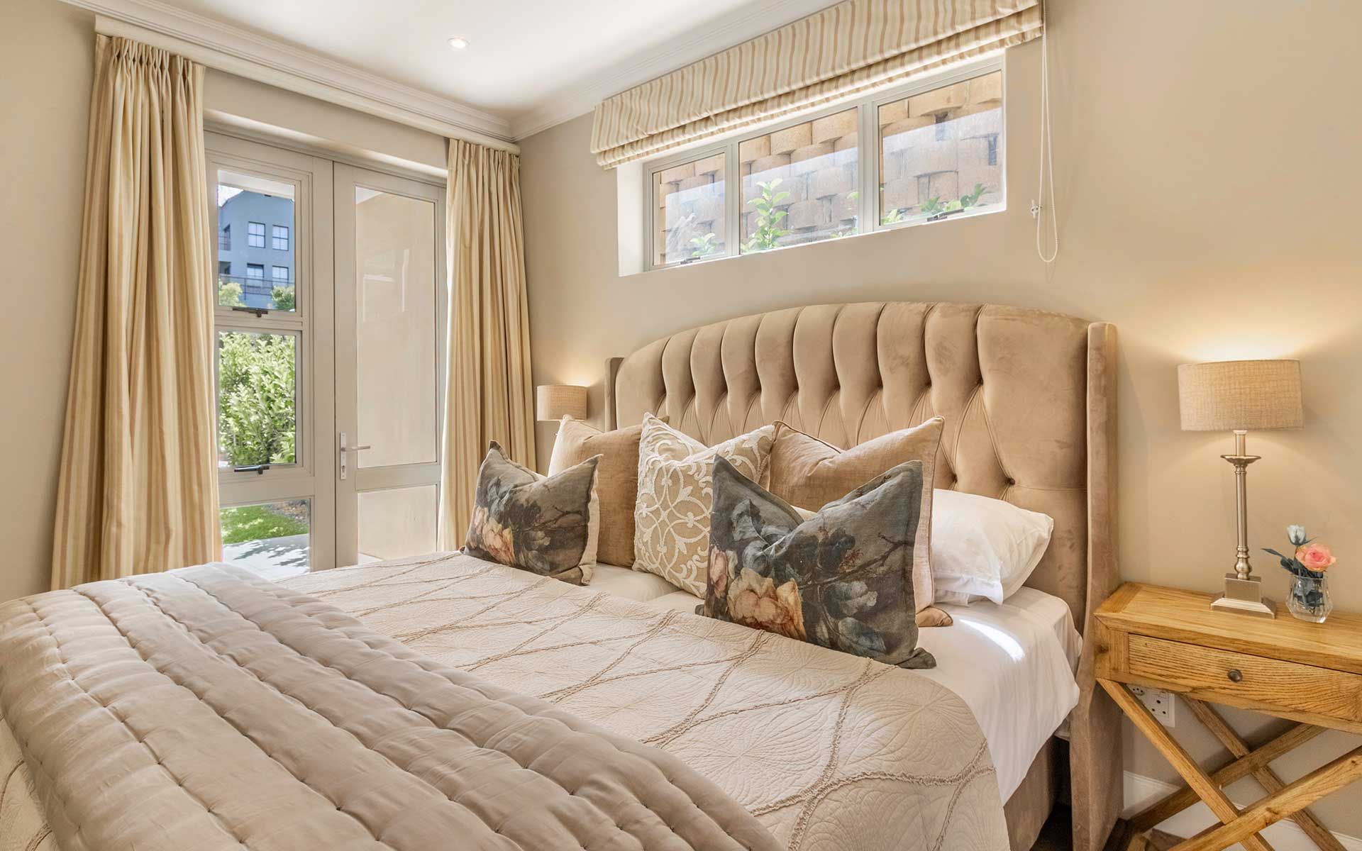 A cozy bedroom in Villa 56 featuring a plush, tufted headboard and elegant bedding. Large windows with beige curtains allow natural light to fill the room, offering views of the lush garden outside. A wooden bedside table with a lamp and flower vase adds a touch of charm.