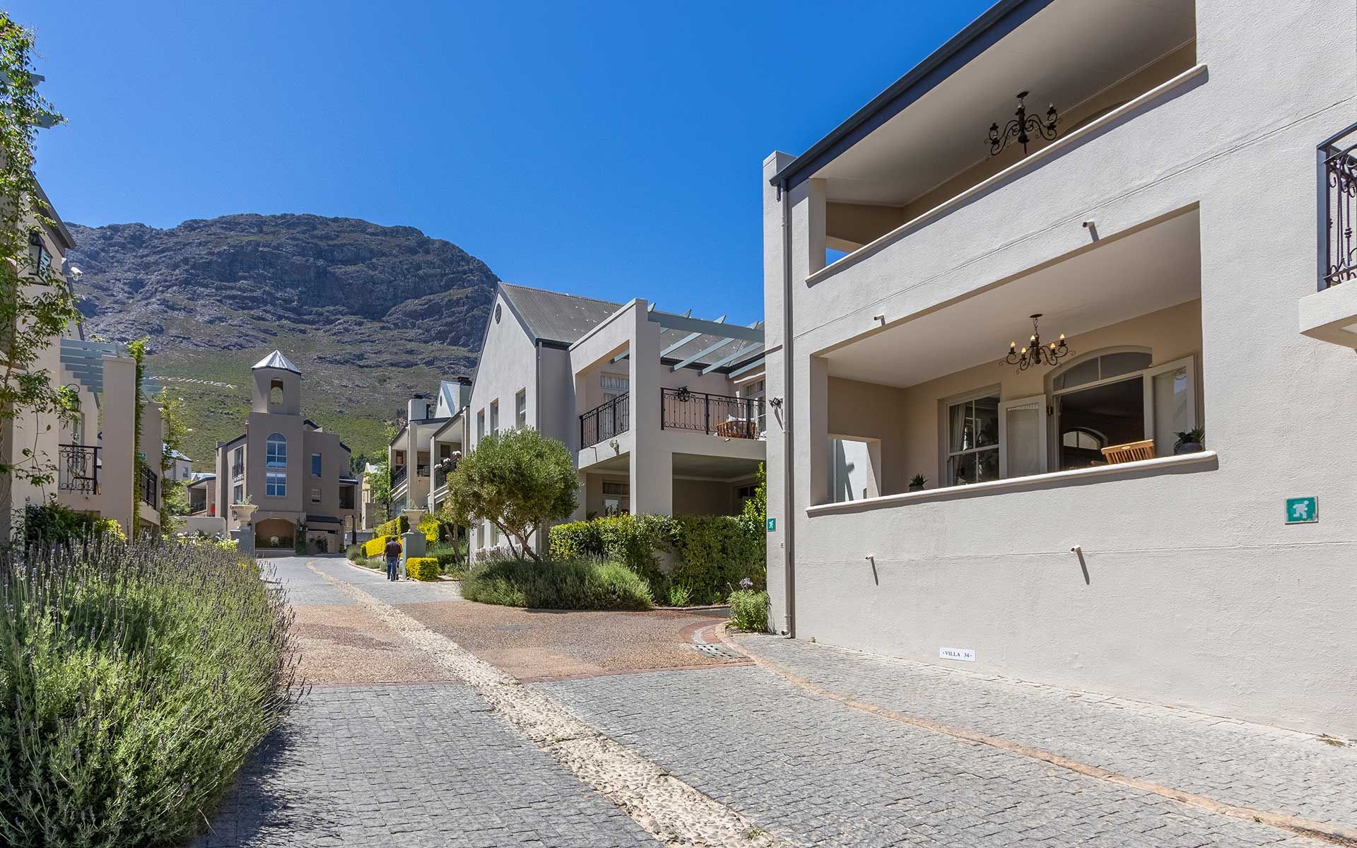 A scenic view of L'Ermitage Village with charming villas lining a cobblestone pathway, set against a backdrop of a majestic mountain under a clear blue sky. The villas feature balconies and lush greenery, creating a tranquil and inviting atmosphere.