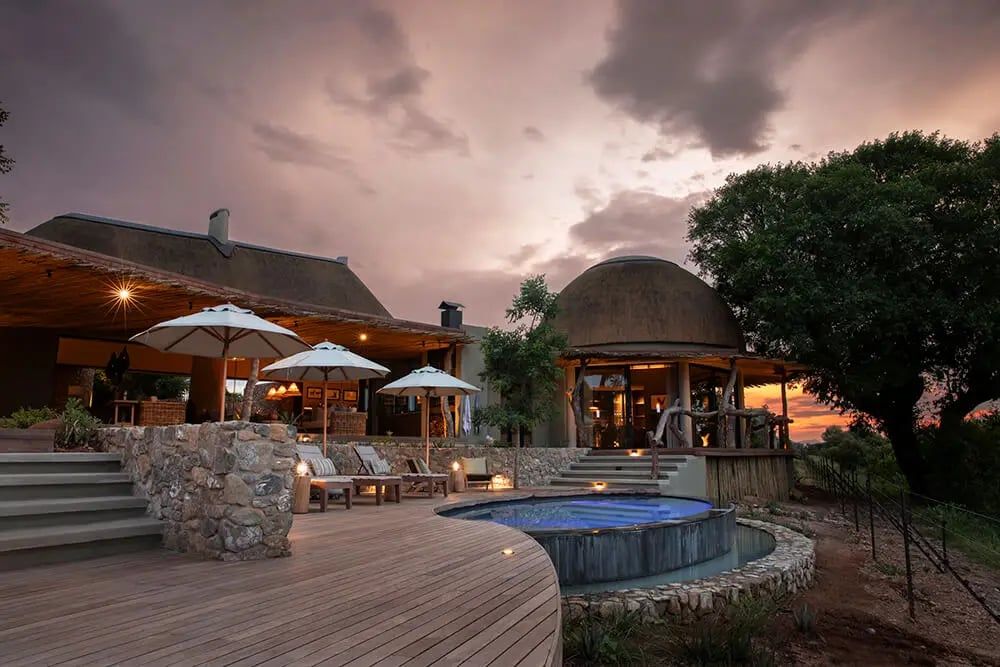 A luxurious lodge with a wooden deck, stone walls, and a circular pool. White umbrellas and lounge chairs offer relaxation under a dramatic sunset sky. The building features curved roofs and large windows, surrounded by lush greenery and a large tree.