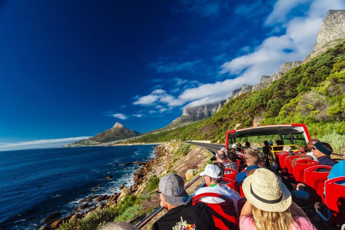 A group of tourists on an open-top red bus admire the scenic coastal view. The road curves along a rocky shoreline with lush green hills and mountains under a clear blue sky. The ocean is calm, dotted with rocks, creating a picturesque travel experience.