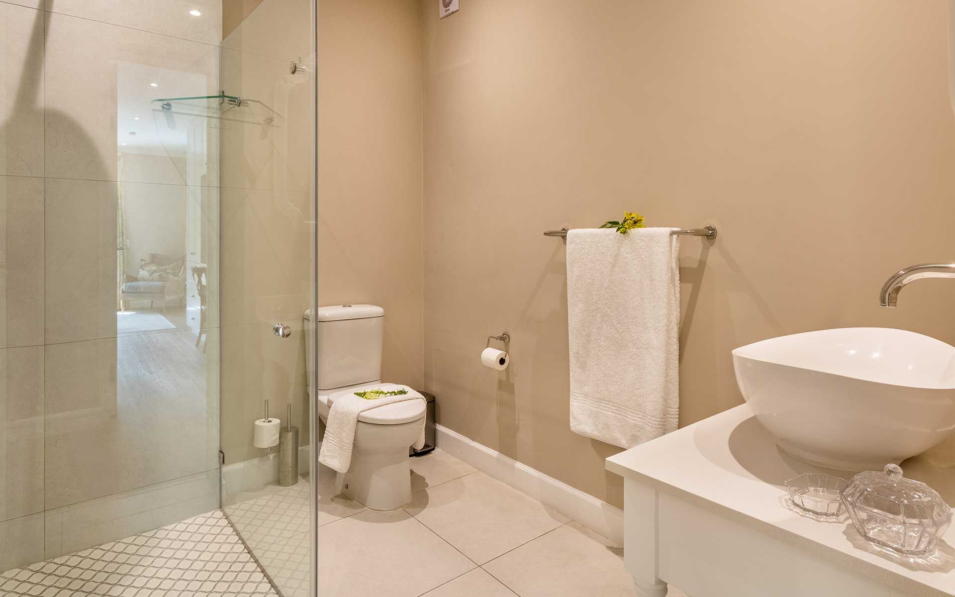 A sophisticated villa bathroom featuring a sleek glass-enclosed shower, modern white sink, and elegant decor. Soft lighting highlights the clean lines and neutral tones, creating a calming atmosphere. A towel rack with fresh towels and subtle floral touches add to the serene ambiance.
