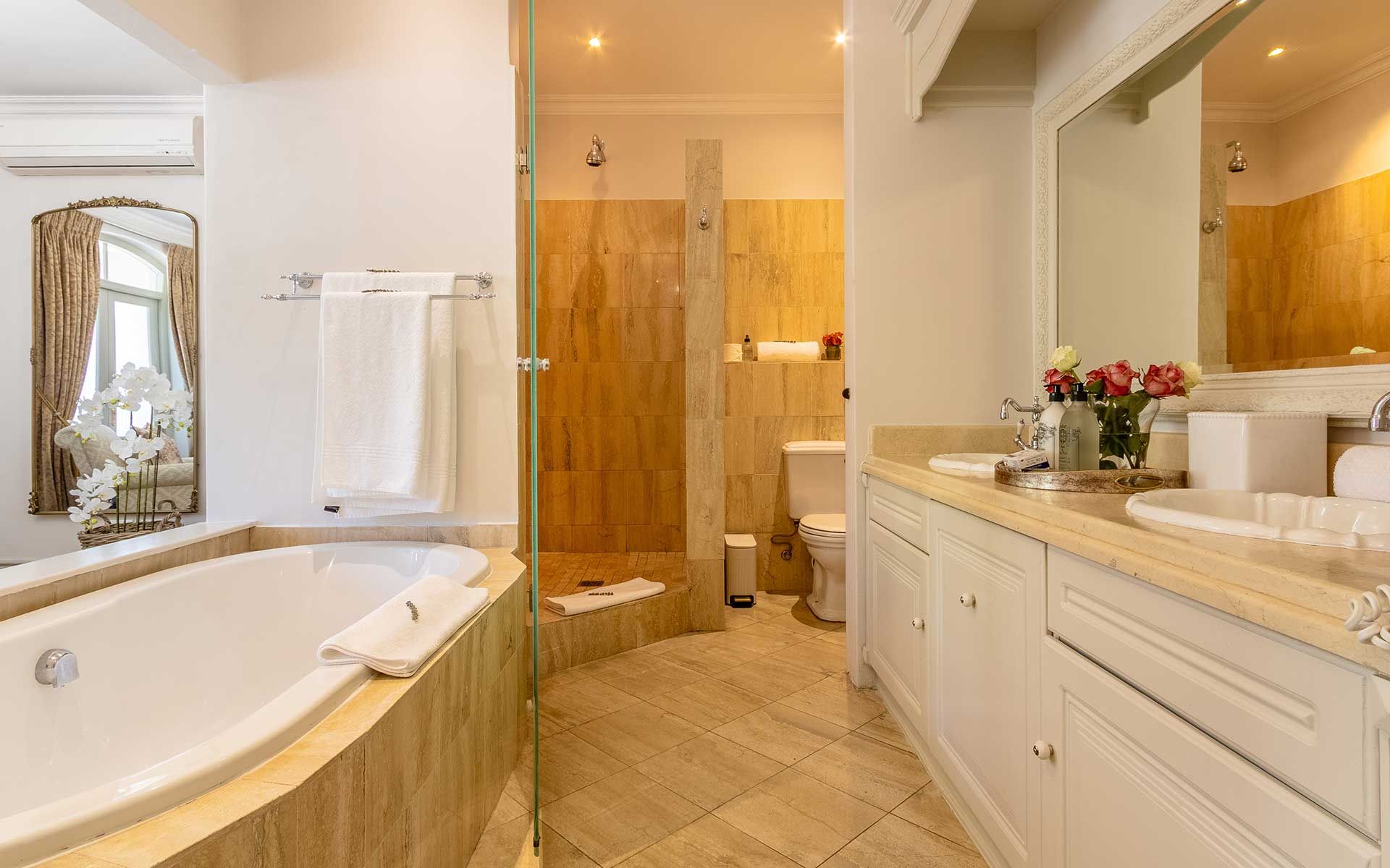 Elegant bathroom in Villa 27 featuring a luxurious bathtub, glass-enclosed shower, and spacious vanity. Warm tones and tasteful decor create a serene ambiance, complemented by fresh flowers and plush towels. Large mirror and natural light enhance the inviting space.
