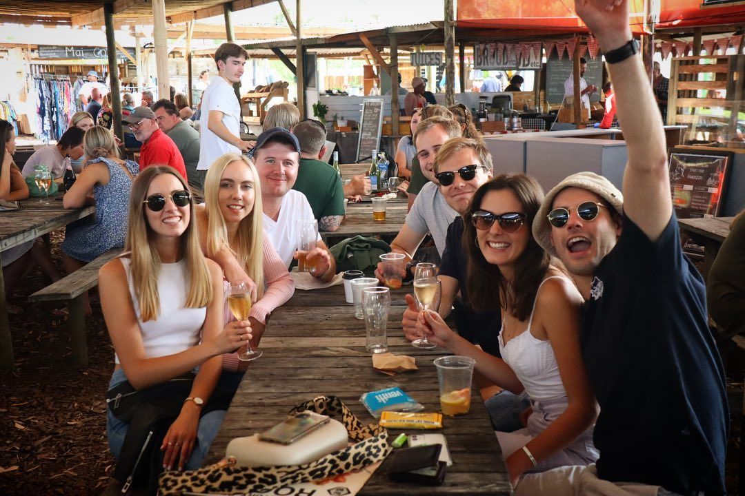 A group of friends enjoying drinks at a long wooden table in the lively Hermanus Country Market. The background shows bustling stalls and cheerful visitors, capturing the warm community atmosphere and vibrant weekend charm that characterizes this local event.