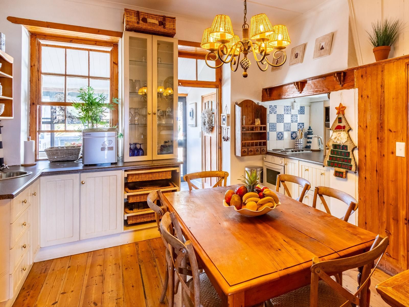 A cozy, rustic dining area with a wooden table surrounded by chairs, adorned with a fruit bowl centerpiece. The adjacent cottage-style kitchen features wooden cabinetry, a small sink, and a window with natural light. Decor includes hanging light fixtures and a festive wall ornament.