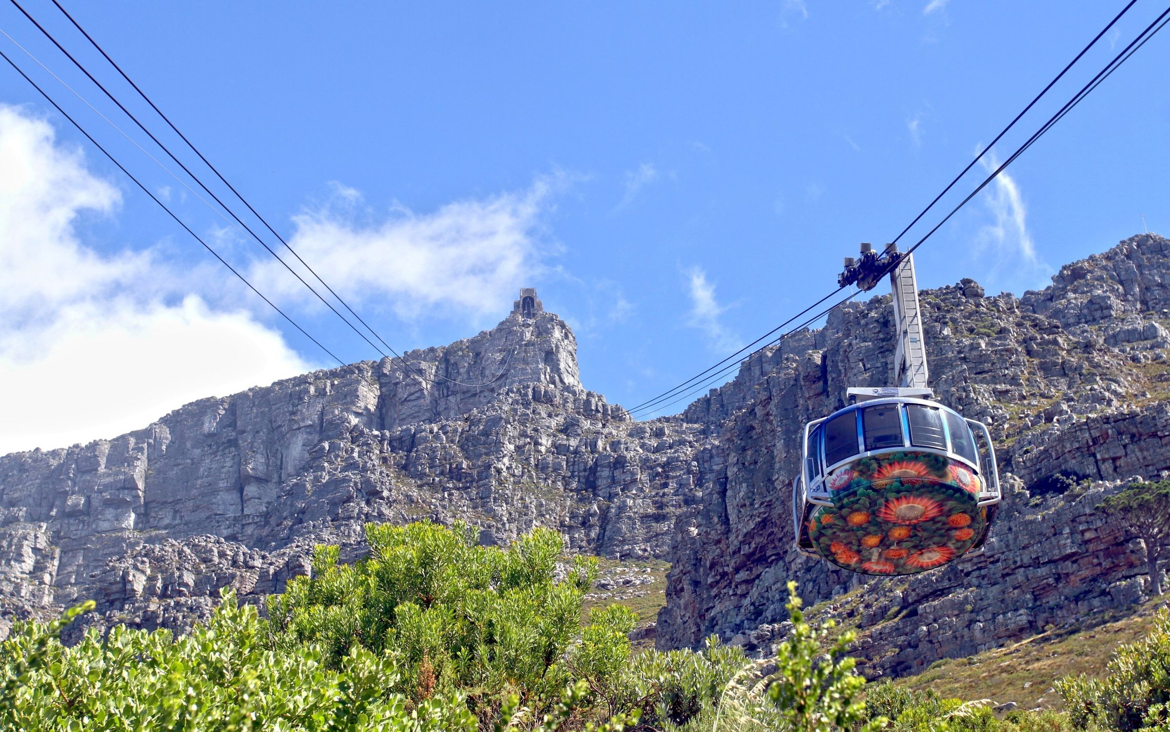 A vibrant cable car ascends towards the flat-topped summit of Table Mountain under a clear blue sky. The rugged cliffs and lush greenery of the fynbos ecosystem are visible, highlighting the natural beauty of this UNESCO World Heritage Site and New Seven Wonders of Nature landmark.