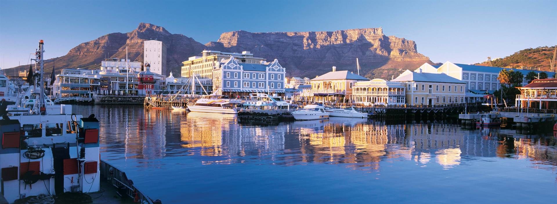 The V&A Waterfront in Cape Town features a lively mix of dining, shopping, and entertainment venues along a bustling harbour. Boats are docked in the calm water, with historic buildings and Table Mountain providing a stunning backdrop under a clear blue sky.