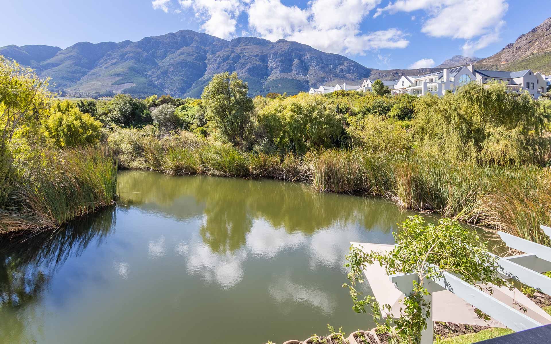 View of tranquil Patrysfontein Lake surrounded by lush greenery, with Mont Rochelle mountains in the background. The scene captures clear blue skies and reflections on the water from the private garden of Villa 31, offering a serene and picturesque setting.
