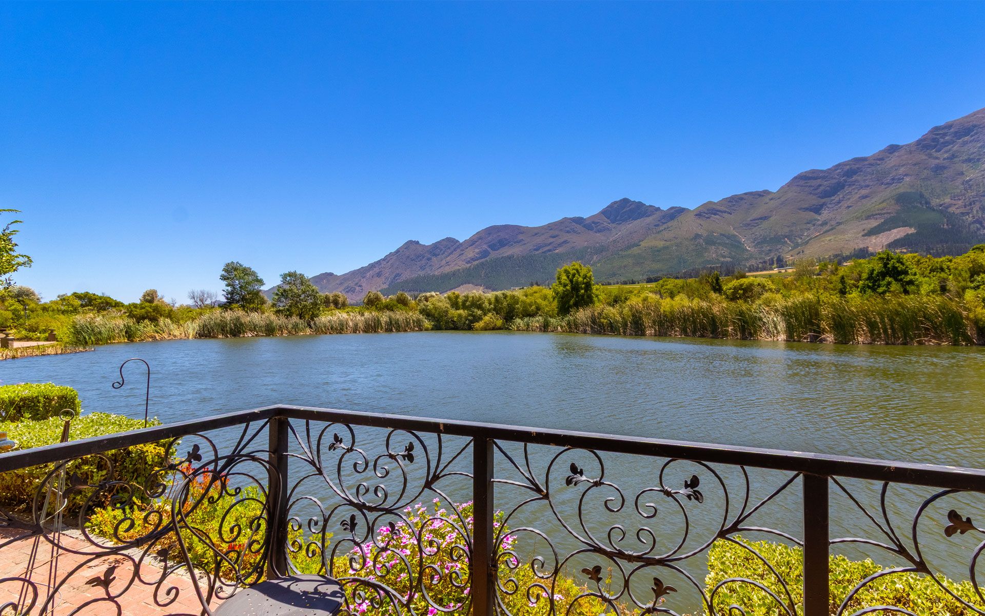 A stunning view from the lakeside patio of Villa 11A at Lermitage, overlooking Patrysfontein Lake with lush greenery and majestic mountains in the background. The ornate railing and vibrant flowers add charm to this tranquil setting in Franschhoek.