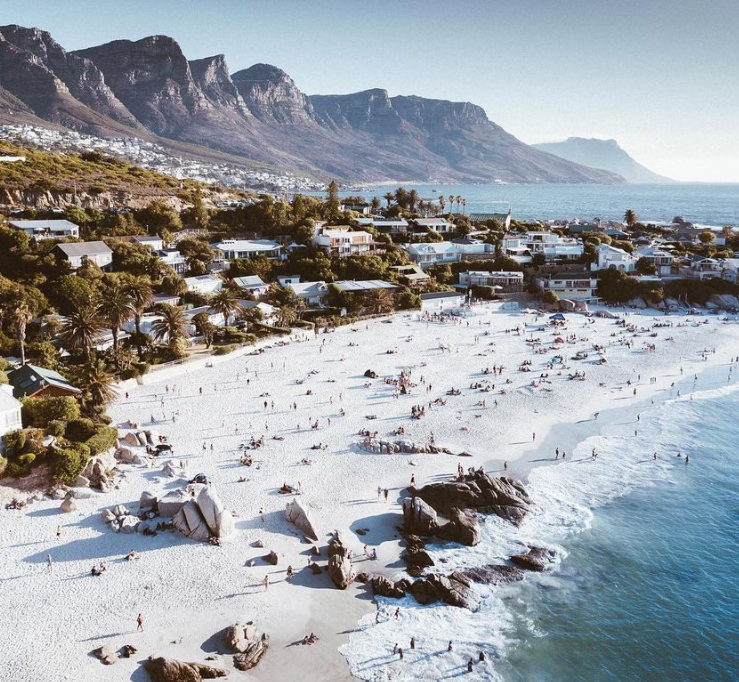 Aerial view of a vibrant Cape Town beach with white sand and turquoise waters. The Twelve Apostles mountain range forms a dramatic backdrop, while people enjoy the sun and surf. The scene captures the contrast between the pristine natural beauty and the nearby urban landscape.