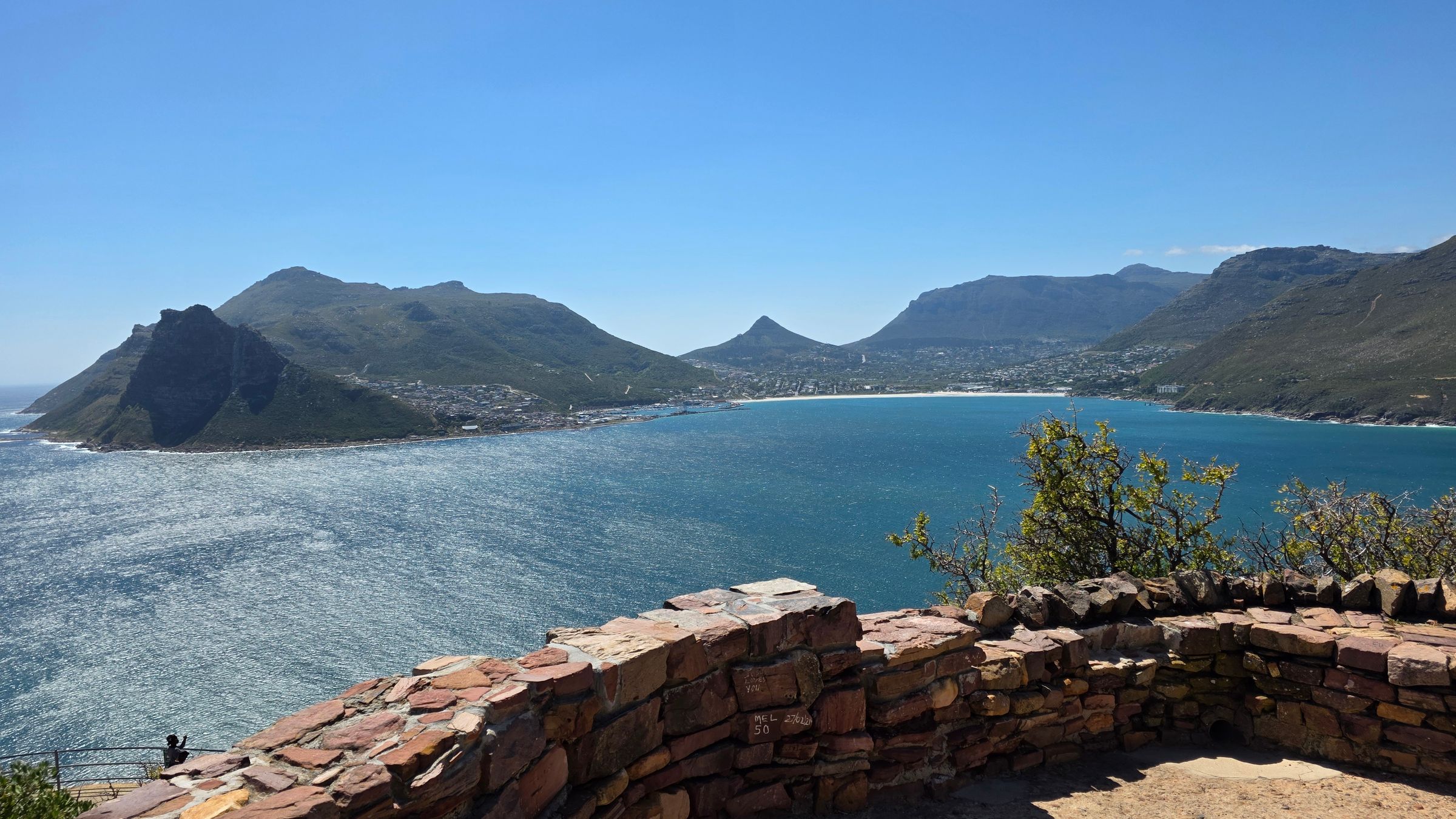 A panoramic view of Hout Bay, showcasing the tranquil blue waters bordered by rugged mountains. In the foreground, a stone wall overlooks the bay, highlighting the charming fishing village atmosphere. The image captures the essence of a serene escape on the Cape Peninsula.