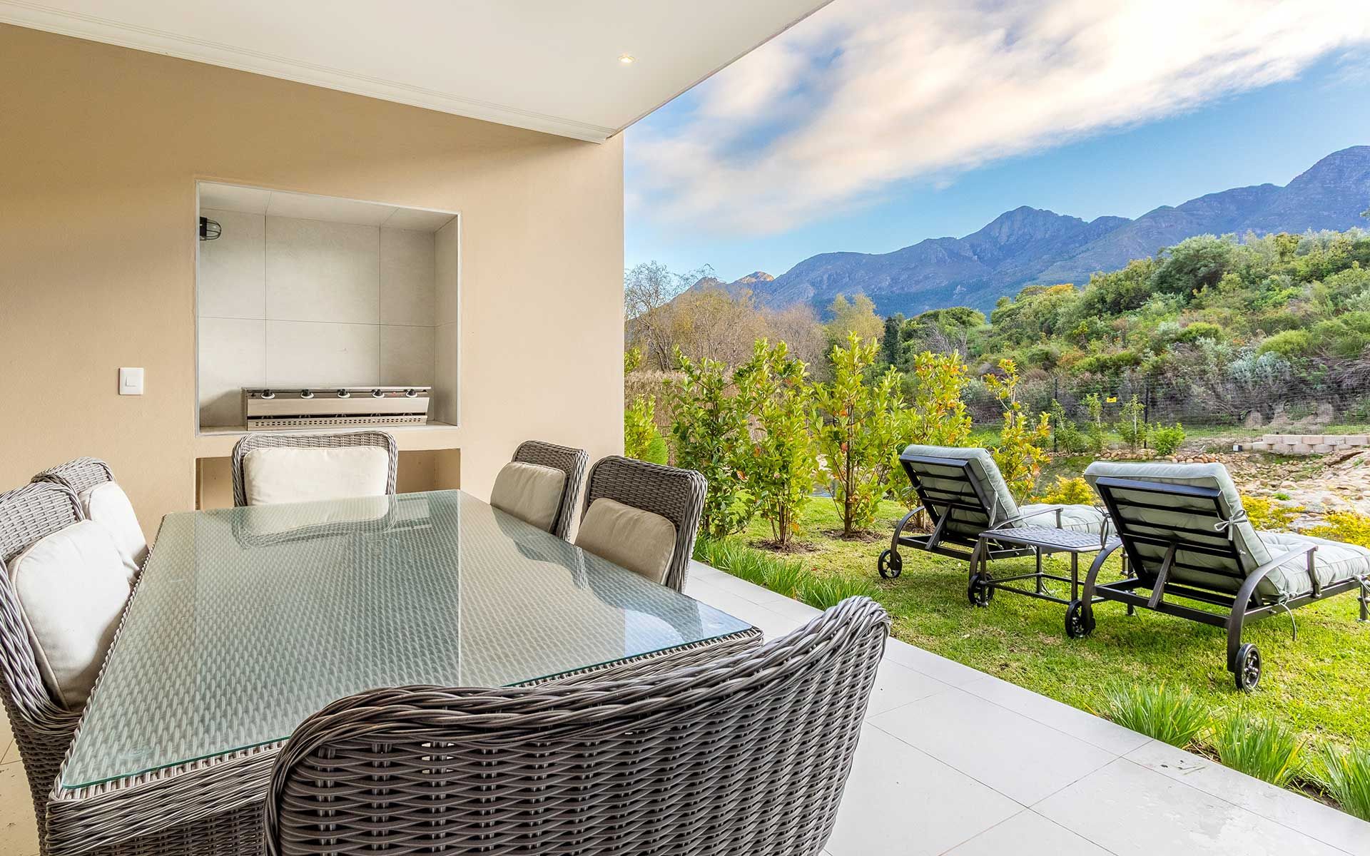 A covered patio with a wicker dining set overlooks lush gardens and mountain views. Two loungers are positioned for relaxation by the greenery, creating a tranquil outdoor space perfect for enjoying the natural surroundings at Villa 56.