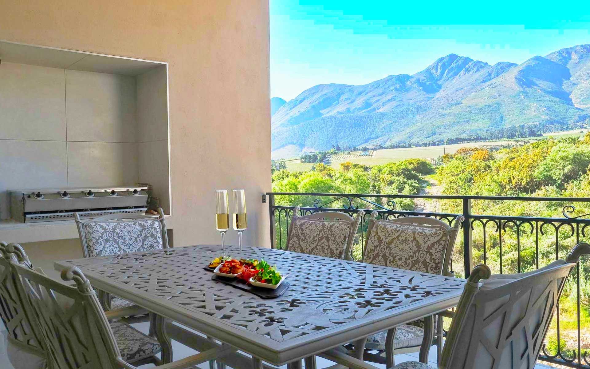 Elegant outdoor dining area of Villa 58 with ornate metal chairs and a table set with appetizers and champagne. The terrace offers a stunning view of Mont Rochelle Nature Reserve and vineyards under a clear blue sky, providing a luxurious and serene atmosphere.
