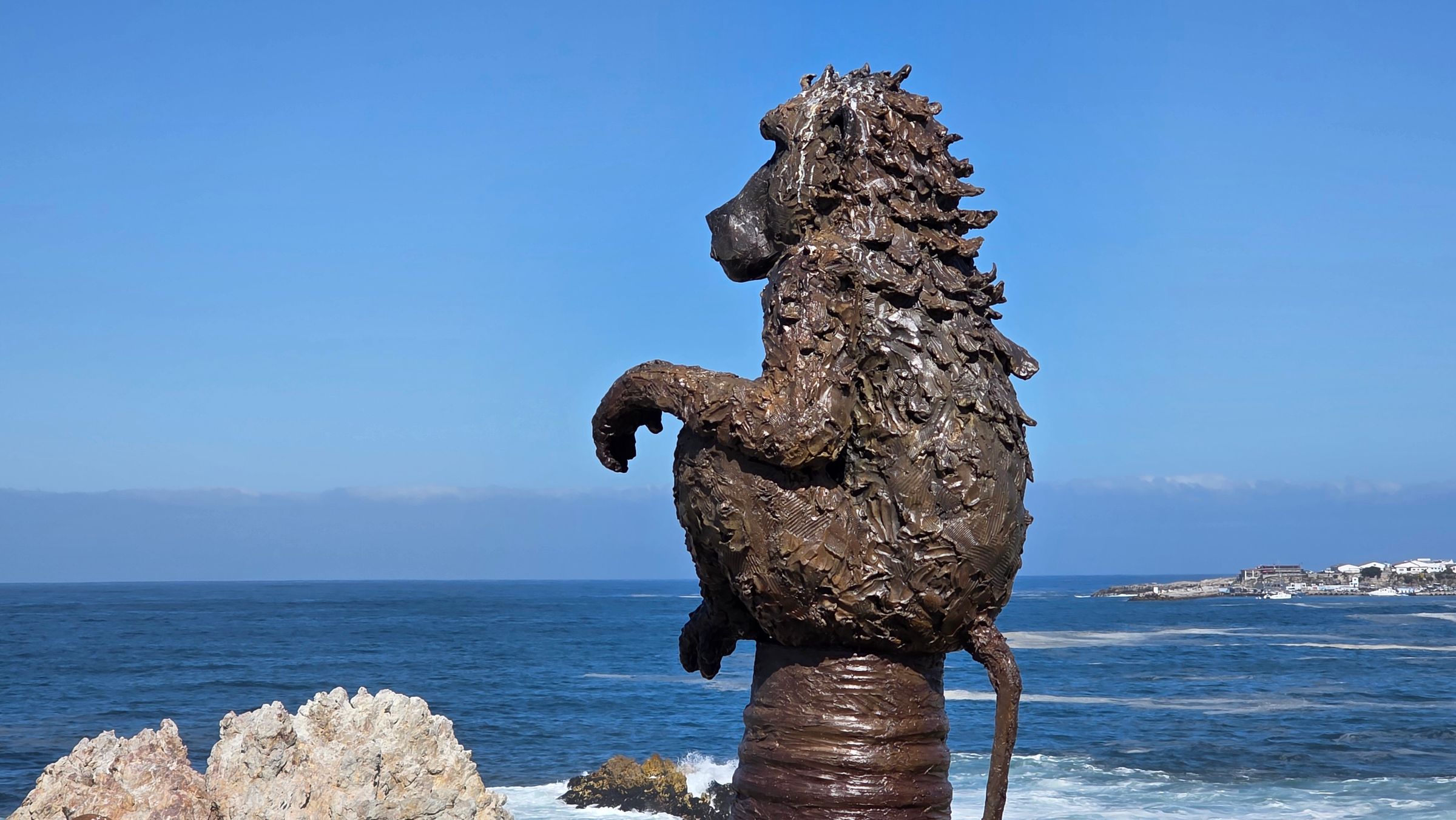 A textured metal sculpture of a baboon sitting on a pedestal, overlooking the ocean. The artwork captures the essence of wildlife in a coastal setting, with a backdrop of blue sea and distant coastline, highlighting Hermanus's blend of art and nature.