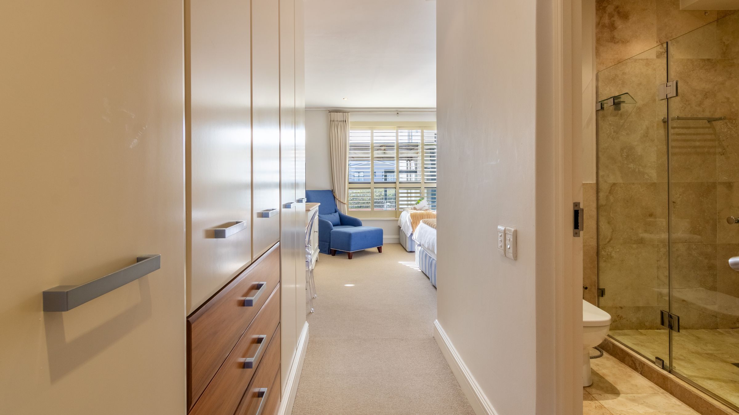 View of a bright bedroom through a hallway with built-in cabinets. The room features a blue armchair by a large window, a bed with light bedding, and a marble-tiled bathroom partially visible on the right. Natural light fills the space, highlighting the neutral tones.