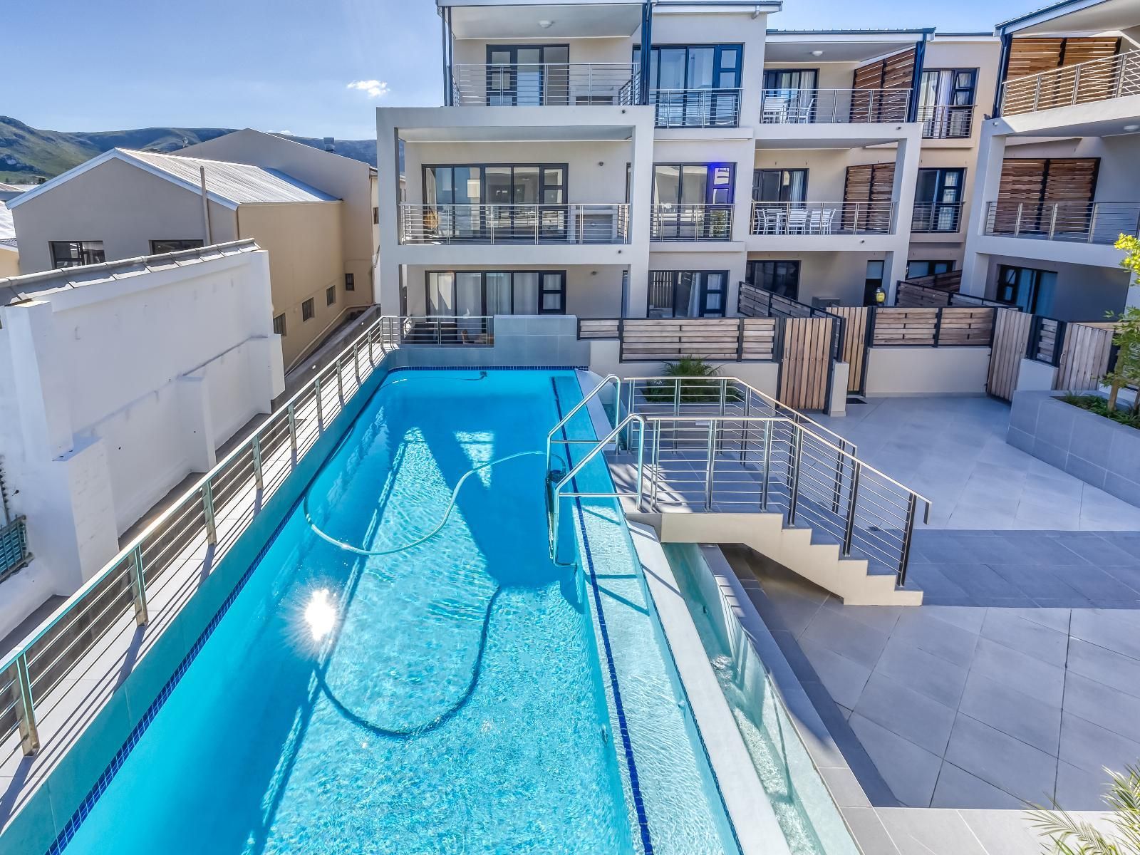 A bright, modern apartment complex featuring multiple balconies overlooking a pristine outdoor swimming pool. Surrounded by mountains, the setting offers a tranquil escape with ample outdoor seating and privacy, embodying luxurious relaxation.