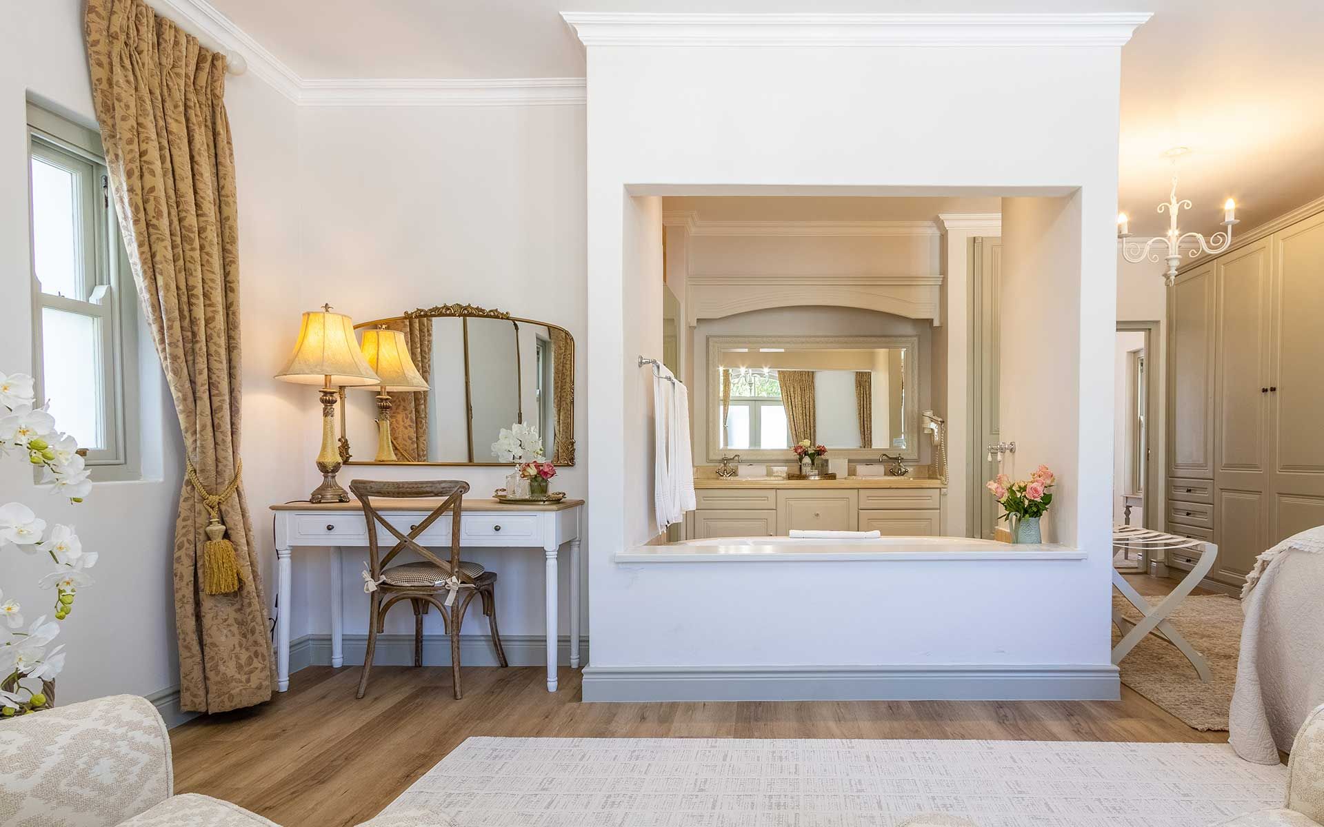A bright, elegant bedroom in Villa 27 with a cozy, inviting atmosphere. Features include a mirrored vanity with a classic chair, warm lamp lighting, and floral decor. The room is styled with soft tones, luxurious drapery, and wooden flooring, creating a tranquil retreat.