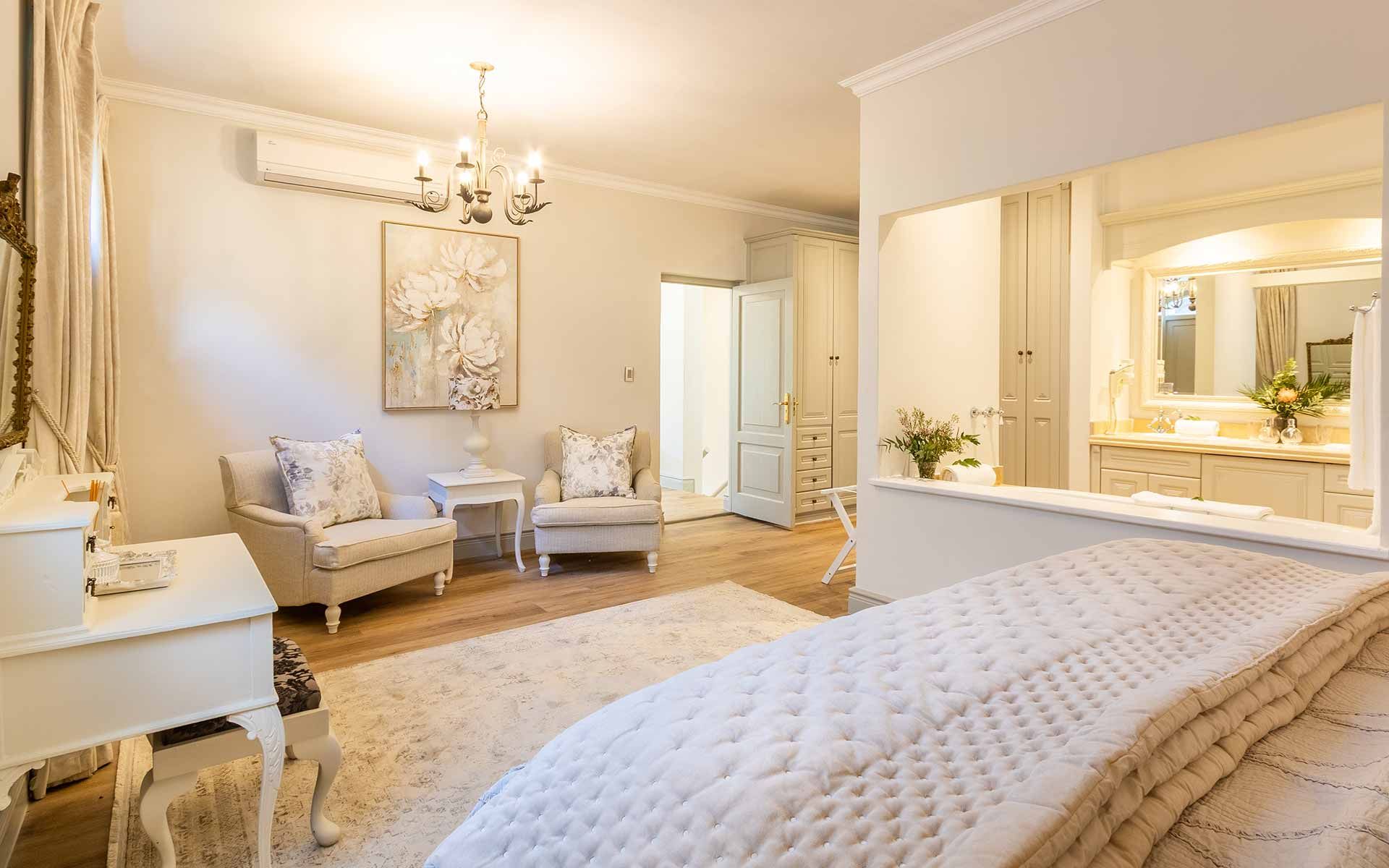 Luxurious bedroom with soft lighting, elegant chandelier, plush seating, and a stylish vanity area. Neutral tones enhance a peaceful ambiance, while decorative art and floral accents add charm. A cozy retreat ideal for relaxation, featuring refined furnishings and ample natural light.