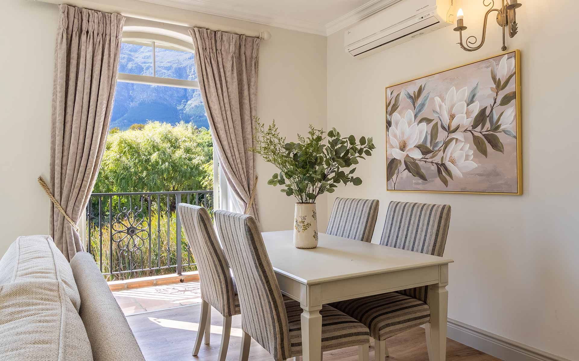 A bright dining area in Villa 31 with a white table, striped chairs, and a vase of greenery. Large window curtains frame views of lush greenery and mountains, enhancing the tranquil atmosphere. Wall art of white flowers adds a decorative touch, and a Juliet balcony is visible outside.