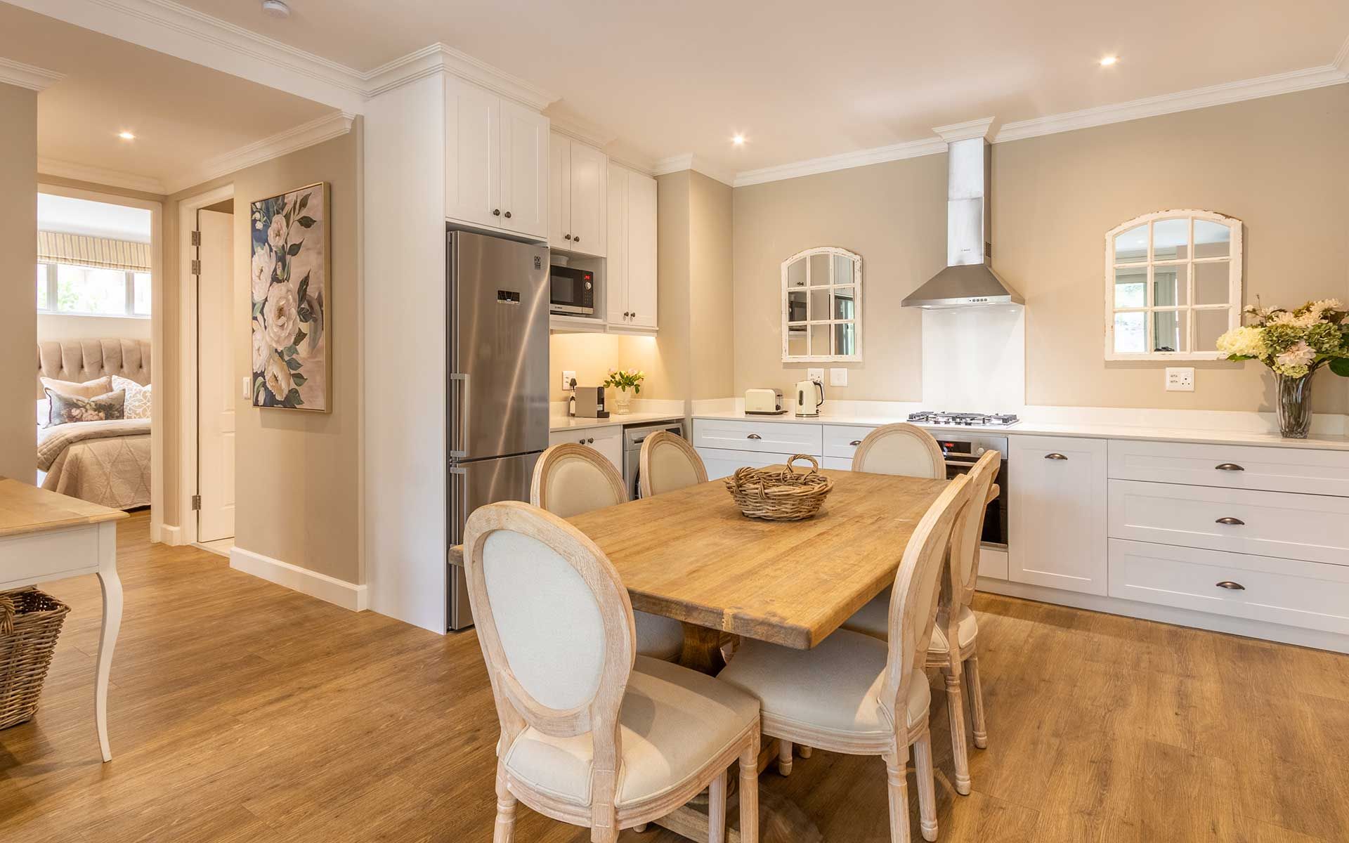 A cozy dining area in Villa 56 featuring a wooden table with elegant chairs, adjacent to a modern kitchen with white cabinetry and stainless steel appliances. A soft-lit interior with floral decor and a glimpse of a comfortable bedroom in the background enhances the tranquil ambiance.