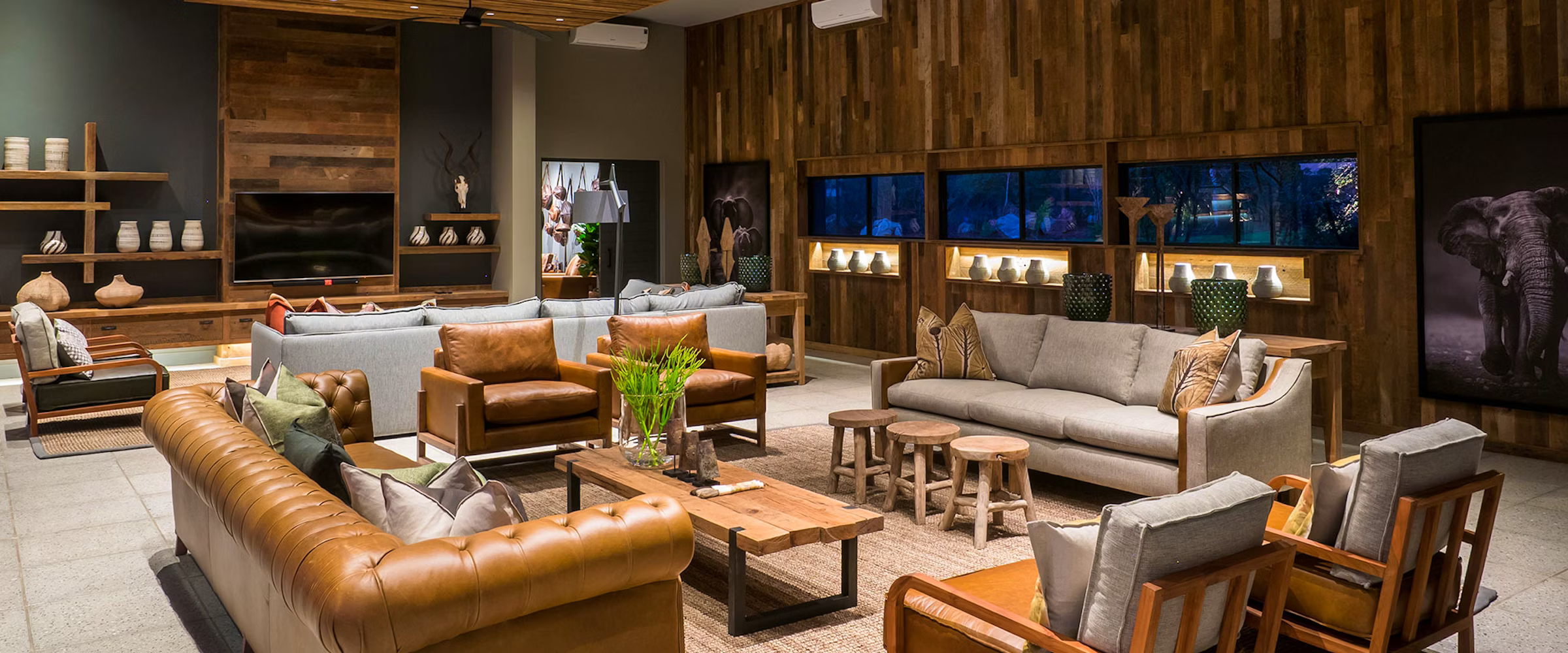 A luxurious lodge living area with leather and fabric seating, wooden coffee tables, and decorative elements. The room features a wooden wall with art and shelves displaying pottery, creating a warm and inviting atmosphere, perfect for relaxation and wildlife viewing.