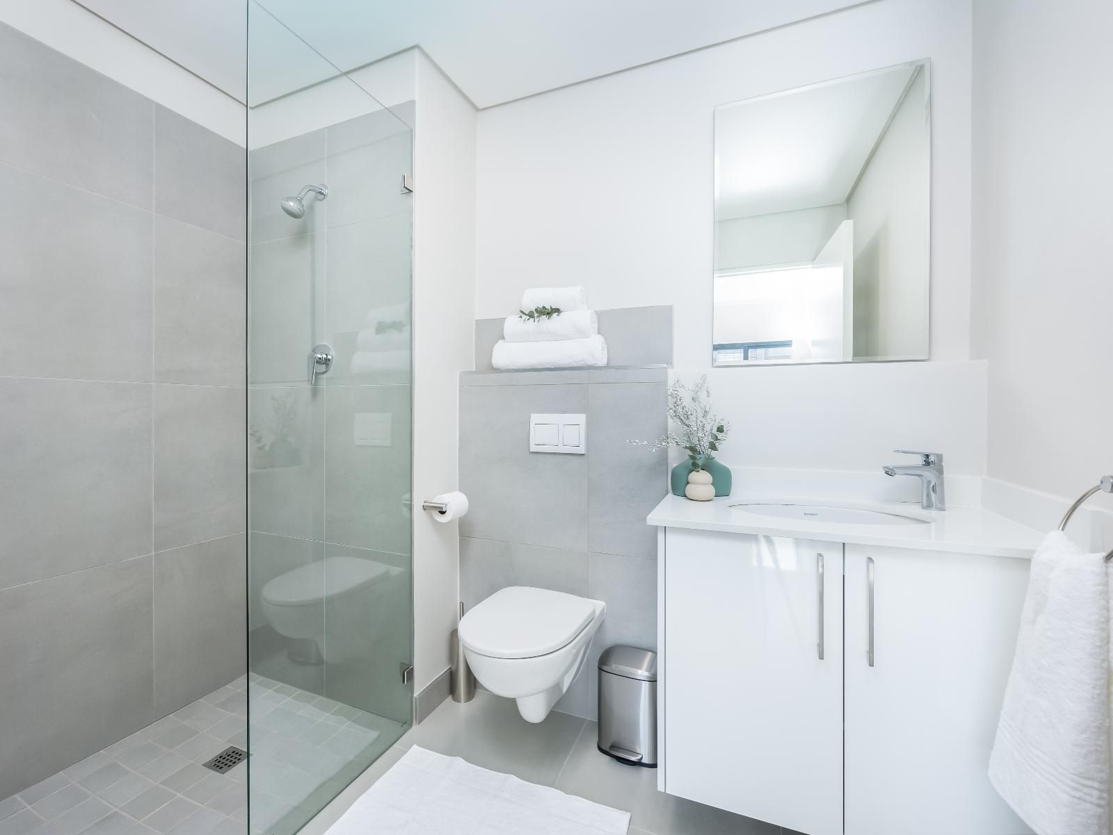 A modern bathroom featuring a glass-enclosed shower, white vanity with sink, large mirror, and toilet. Towels are neatly arranged, and a small plant adds a touch of greenery, enhancing the serene atmosphere with minimalist design and bright lighting.