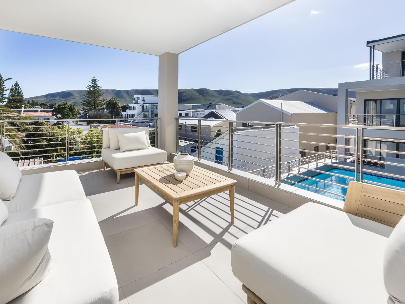 A modern apartment balcony featuring comfortable white cushioned seating and a wooden coffee table. The space overlooks a residential area with a pool and offers scenic views of distant hills and clear blue skies, enhancing the sense of tranquility.