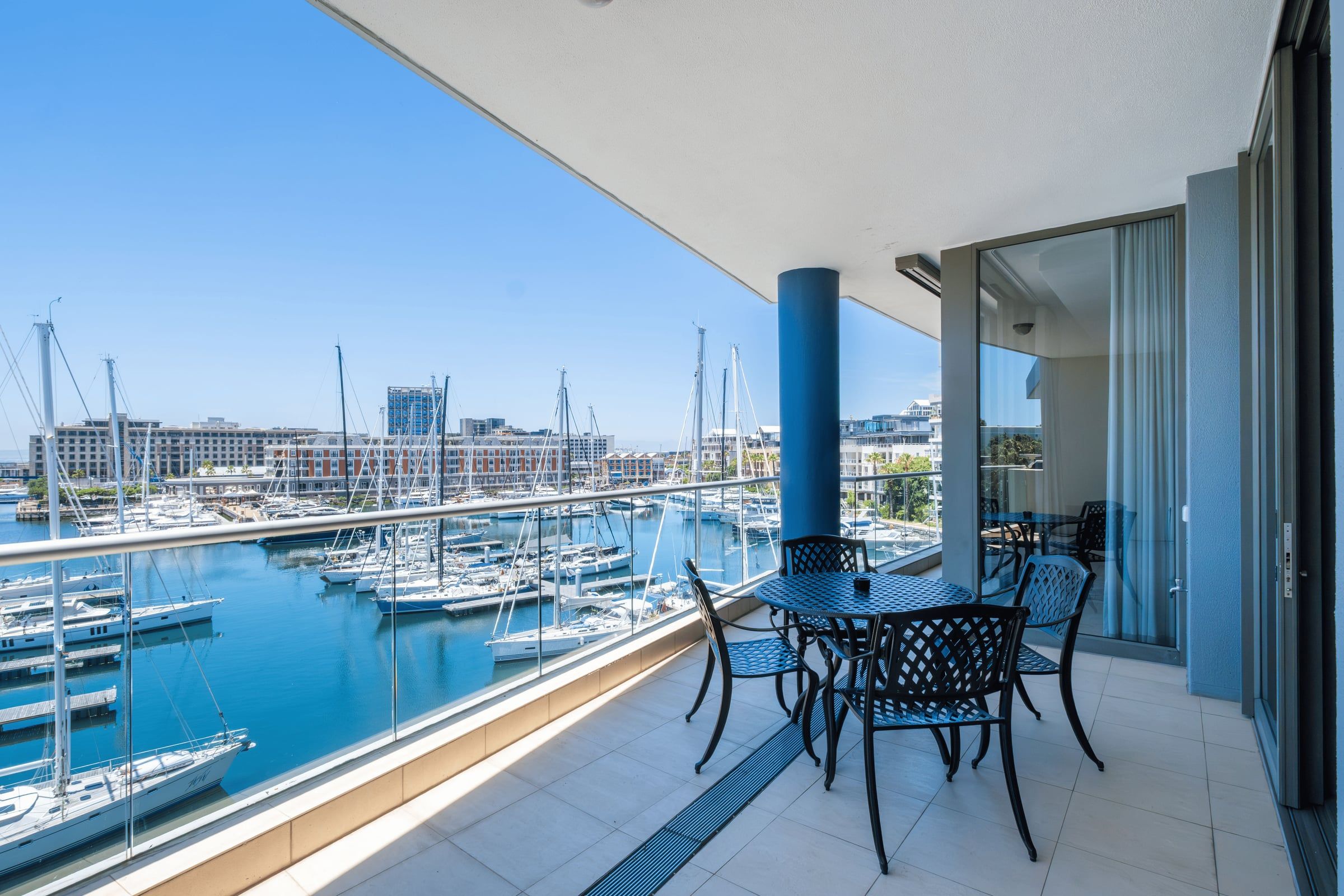 A spacious balcony overlooking a marina filled with sailboats under a clear blue sky. The balcony features a black metal table and chairs set on a tiled floor. Modern buildings are visible in the background, enhancing the scenic coastal ambiance.