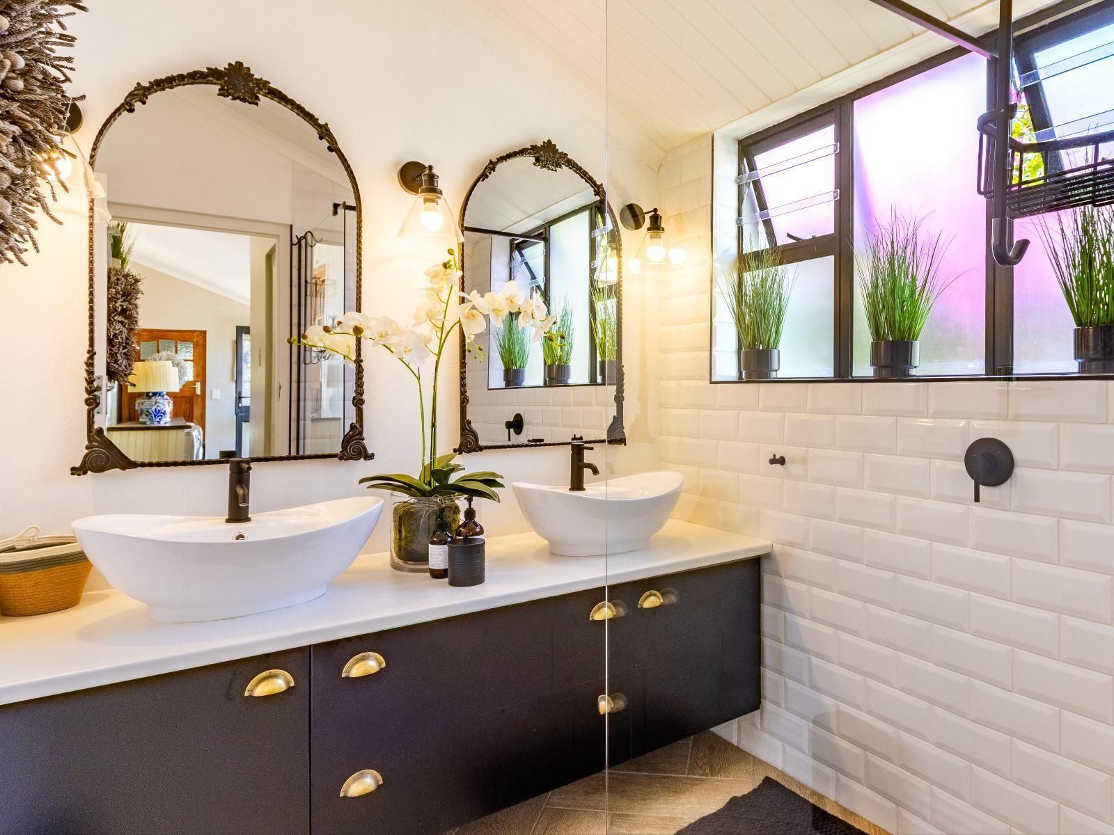A luxurious bathroom featuring a dual vanity with elegant vessel sinks, ornate mirrors, and modern lighting. The space includes frosted windows with potted plants, adding a touch of greenery to the sleek white and black decor.