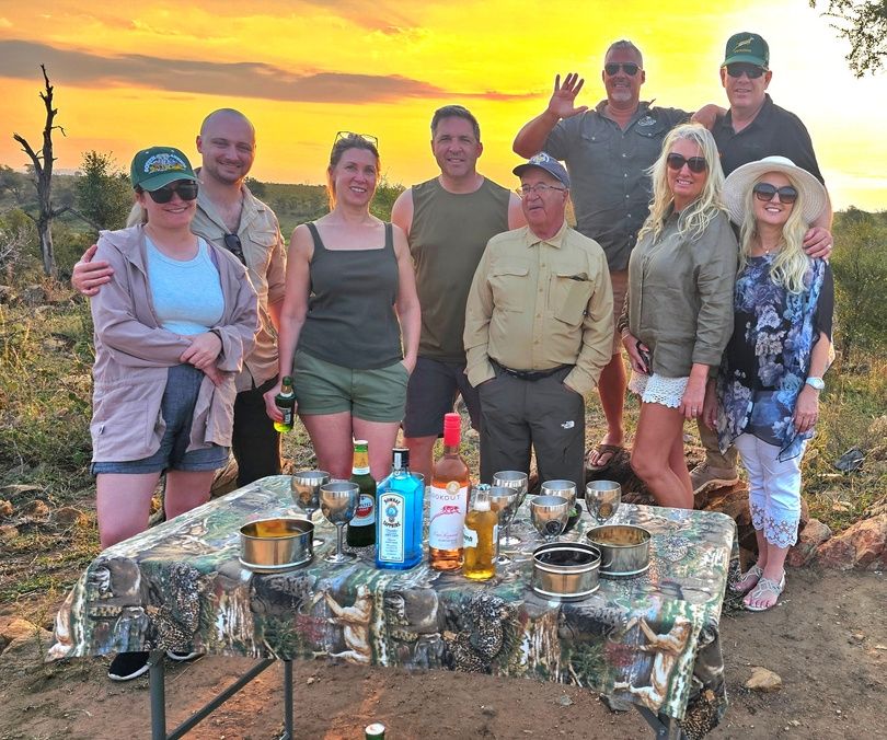 A group of nine people posing outdoors with a sunset backdrop. They stand around a table with drinks and glasses. The setting appears to be a natural environment with trees and a clear sky. The group is casually dressed, and some are smiling at the camera.