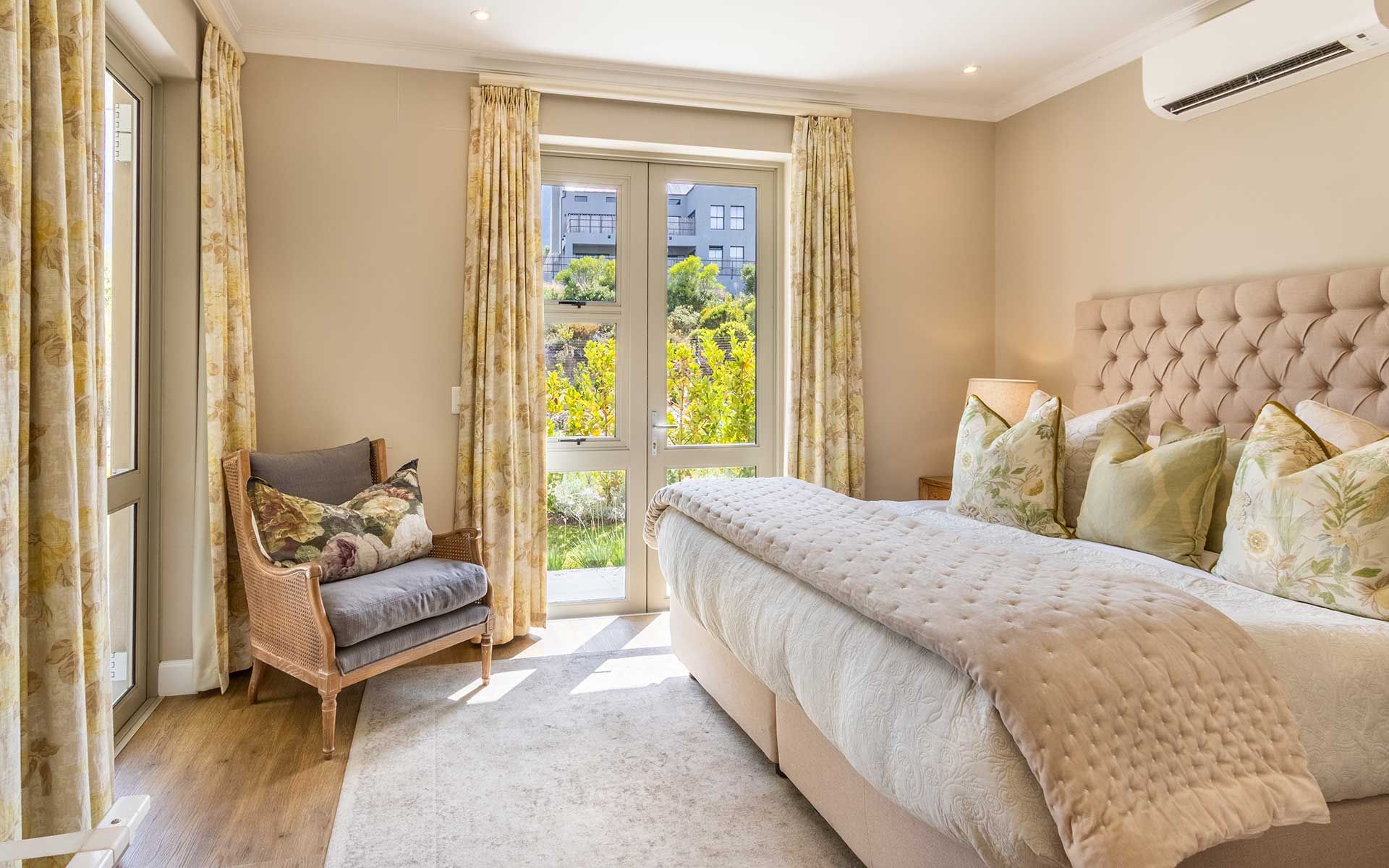 A luxurious bedroom featuring a plush king-size bed with floral pillows, soft beige bedding, and a tufted headboard. Sunlight streams through large windows with floral curtains, highlighting a cozy chair and the serene garden view outside.