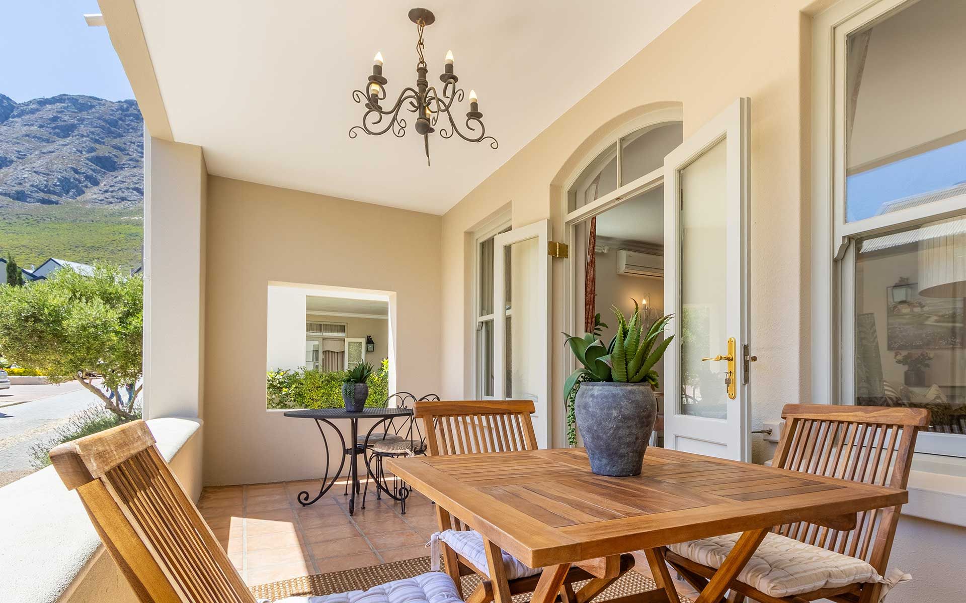 A charming balcony at Villa 27 with wooden furniture and lush potted plants. Overlooks a tranquil village and mountainous landscape, offering a serene spot for relaxation. Features a decorative chandelier and open French doors leading inside, bathed in natural light.