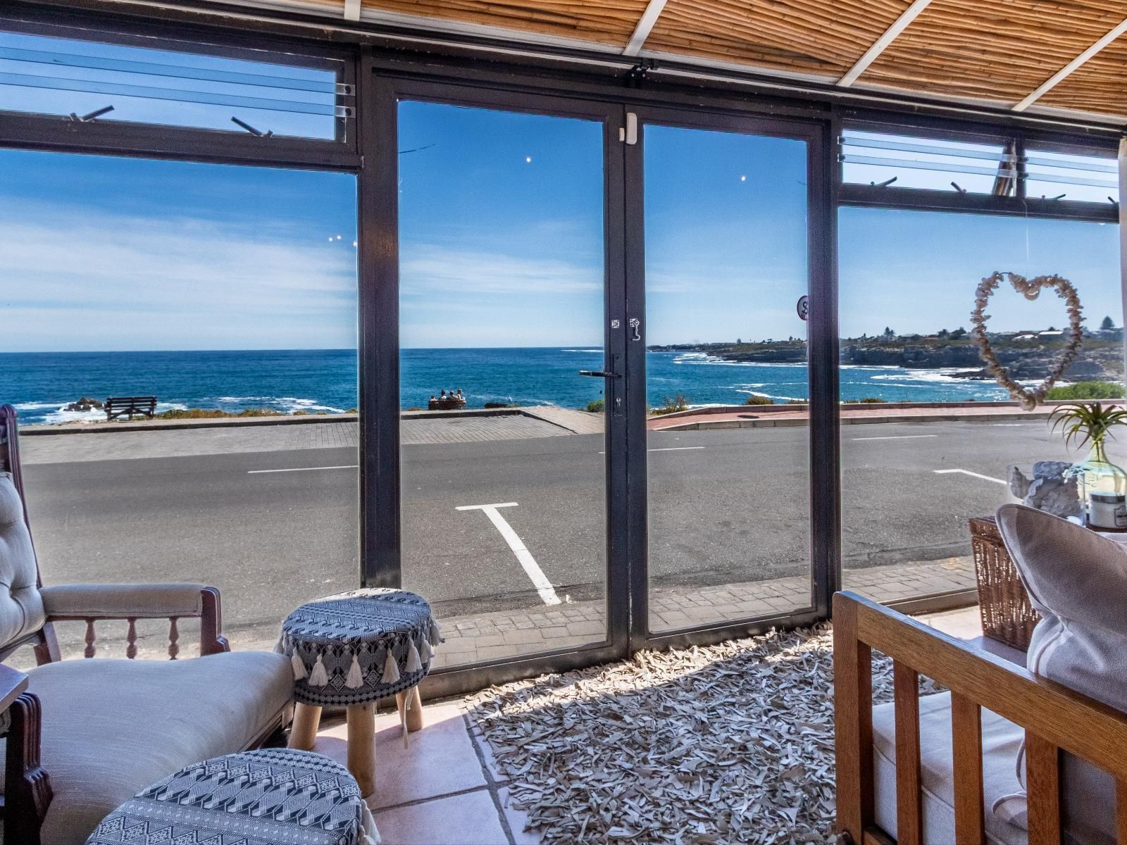 A bright sunroom with large glass doors offers stunning ocean views. Cozy seating includes cushioned chairs and stools, facing the sea. The decor is light and inviting, featuring a natural woven rug and a heart-shaped ornament. Ideal for whale watching and enjoying sunsets.