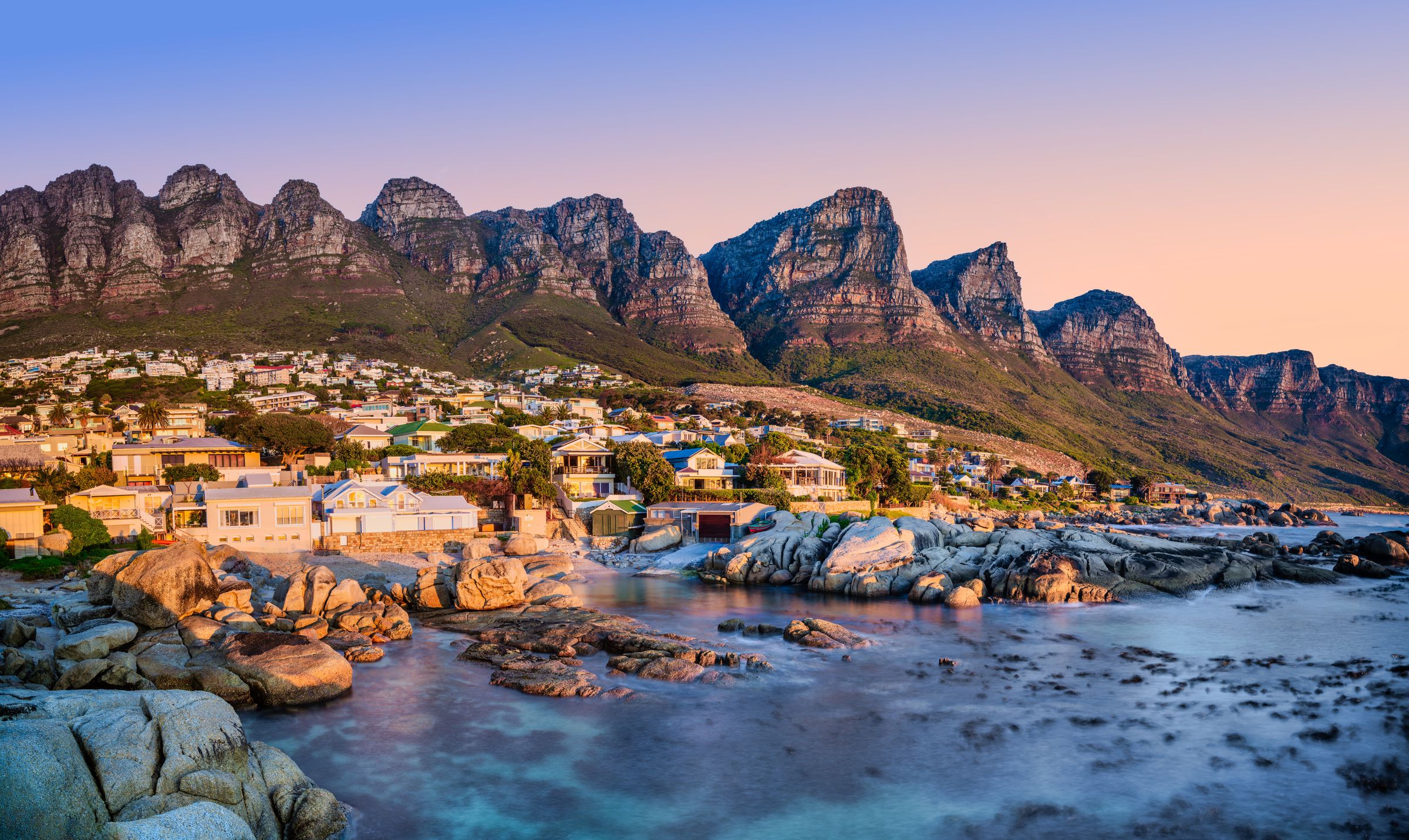 A stunning view of Camps Bay at sunset, with the Twelve Apostles mountain range in the background. The foreground features rocky shores and turquoise waters, with a vibrant beachside neighborhood nestled against the dramatic scenery, reflecting the golden hues of the setting sun.