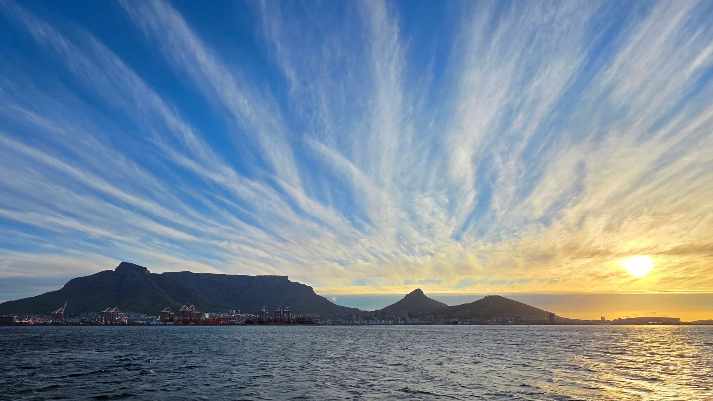 Panoramic view of Table Mountain's flat-topped summit against a dramatic sunset sky in Cape Town. The iconic skyline features the mountain's silhouette, the ocean in the foreground, and vibrant clouds, highlighting its status as a UNESCO World Heritage Site and a New Seven Wonders of Nature.