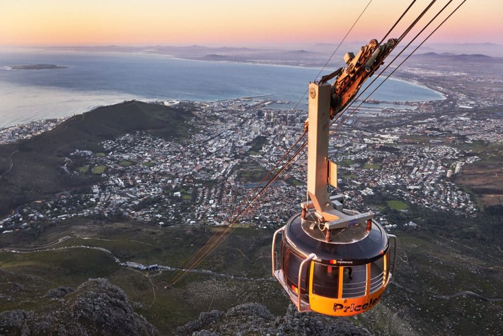 A cable car ascends above Cape Town, offering a panoramic view of the city, surrounding mountains, and the ocean at sunset. The horizon is painted in soft pink and orange hues, highlighting the scenic landscape and urban sprawl below.