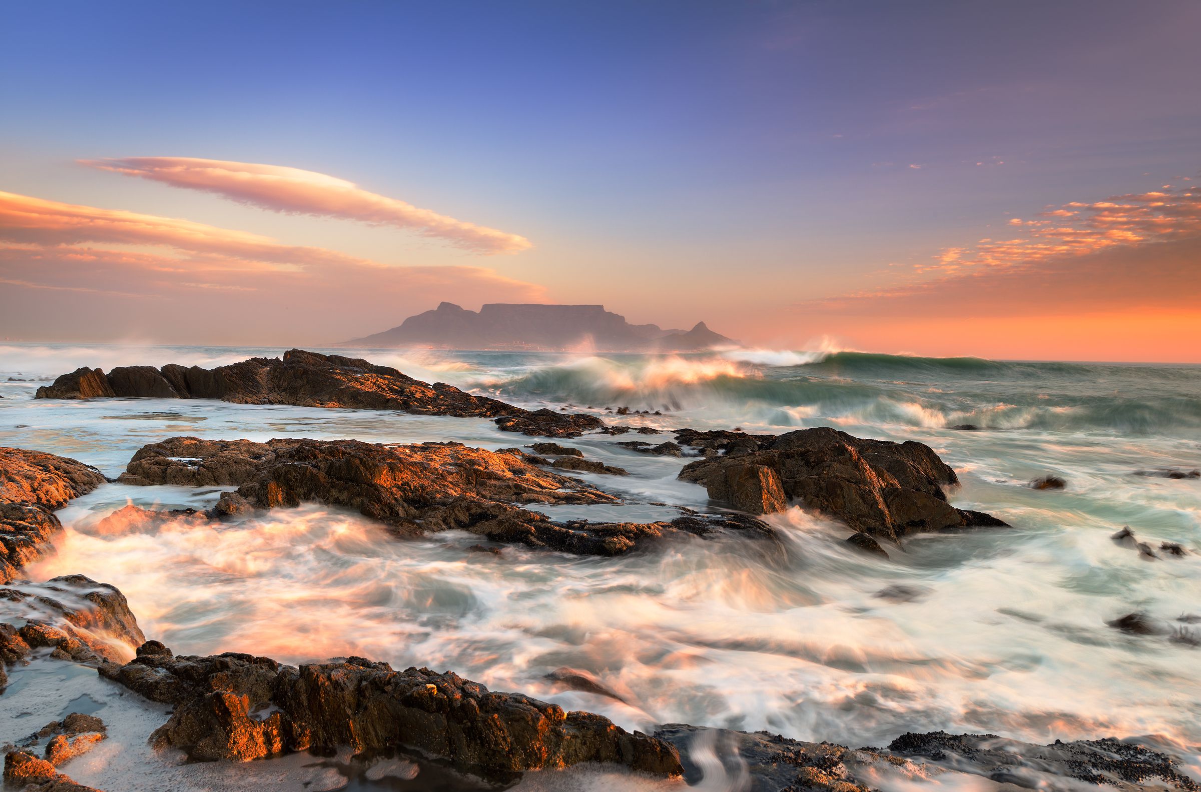 A stunning view of Table Mountain from the rocky coast at sunrise. The flat-topped summit stands majestically against a colorful sky, with waves crashing onto the rocks in the foreground. The scene captures the mountain's iconic presence and the surrounding ocean's dynamic beauty.