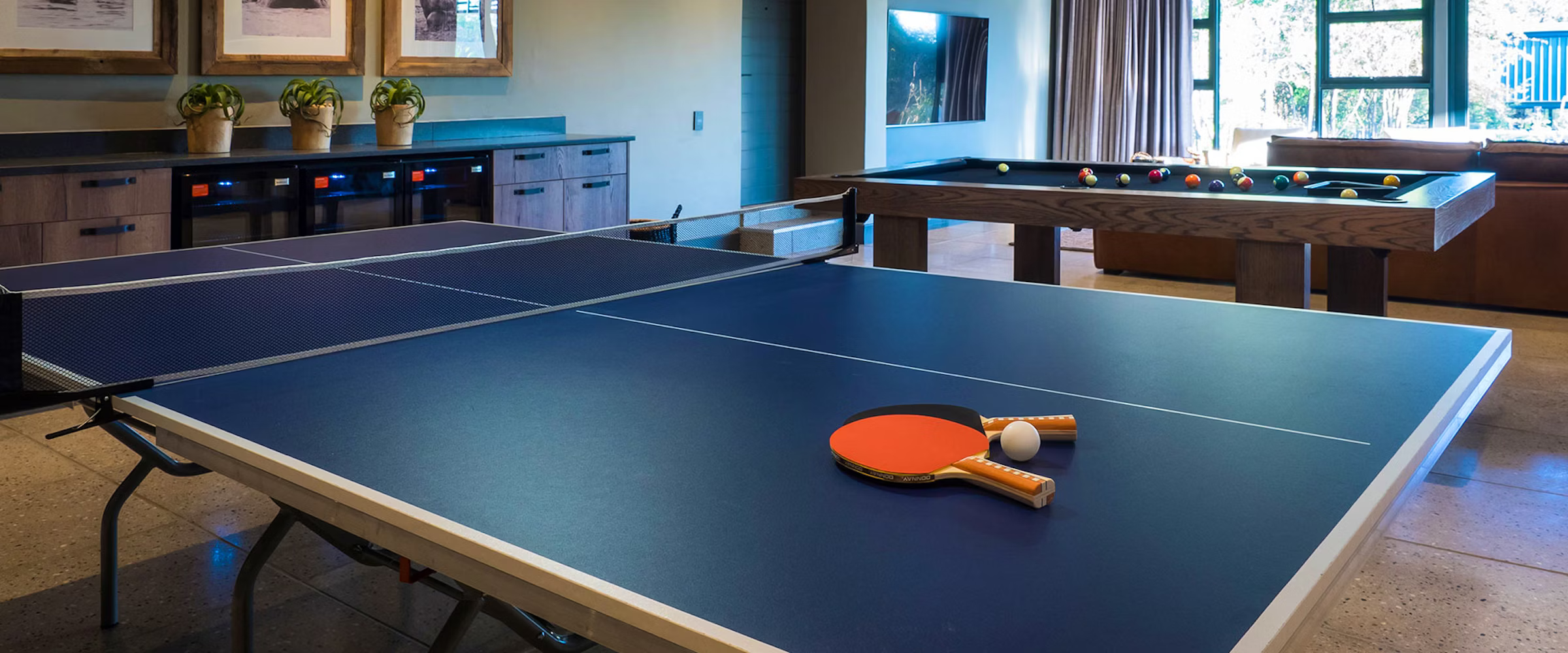 A luxury lodge game room featuring a ping pong table with paddles and ball, alongside a billiards table. The space is elegantly designed with modern decor, framed artwork, and large windows allowing natural light, enhancing the upscale safari experience.